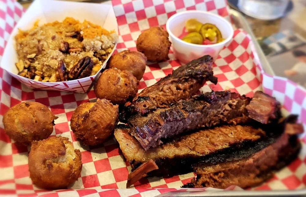 Barbecue ribs, fried sweet potato balls, baked macaroni and cheese, and jalapeño pickles on a red checkered paper-lined tray.