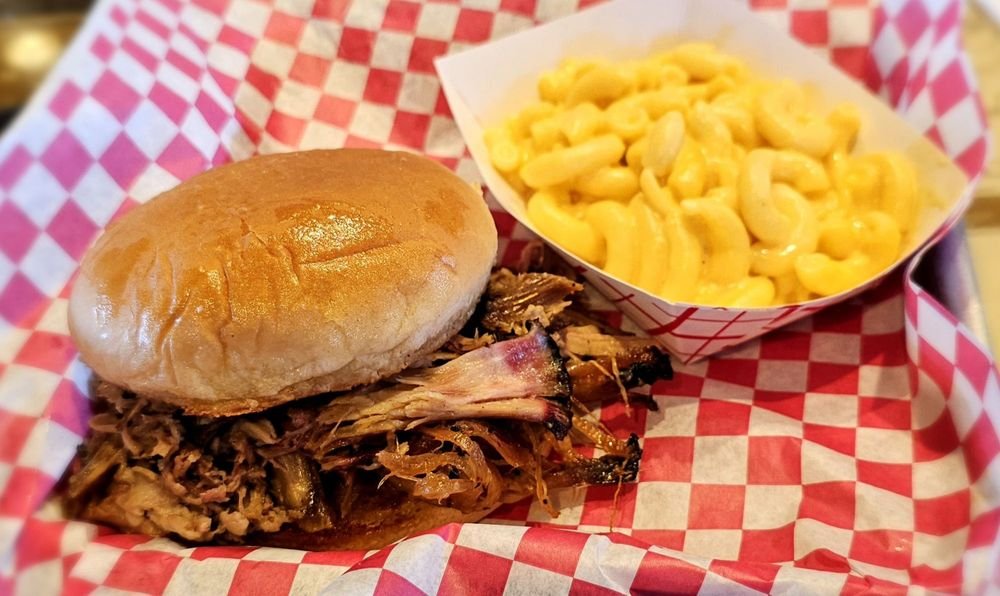 Pulled pork barbecue sandwich with a bun, served with a side of macaroni and cheese in a red and white checkered paper tray.