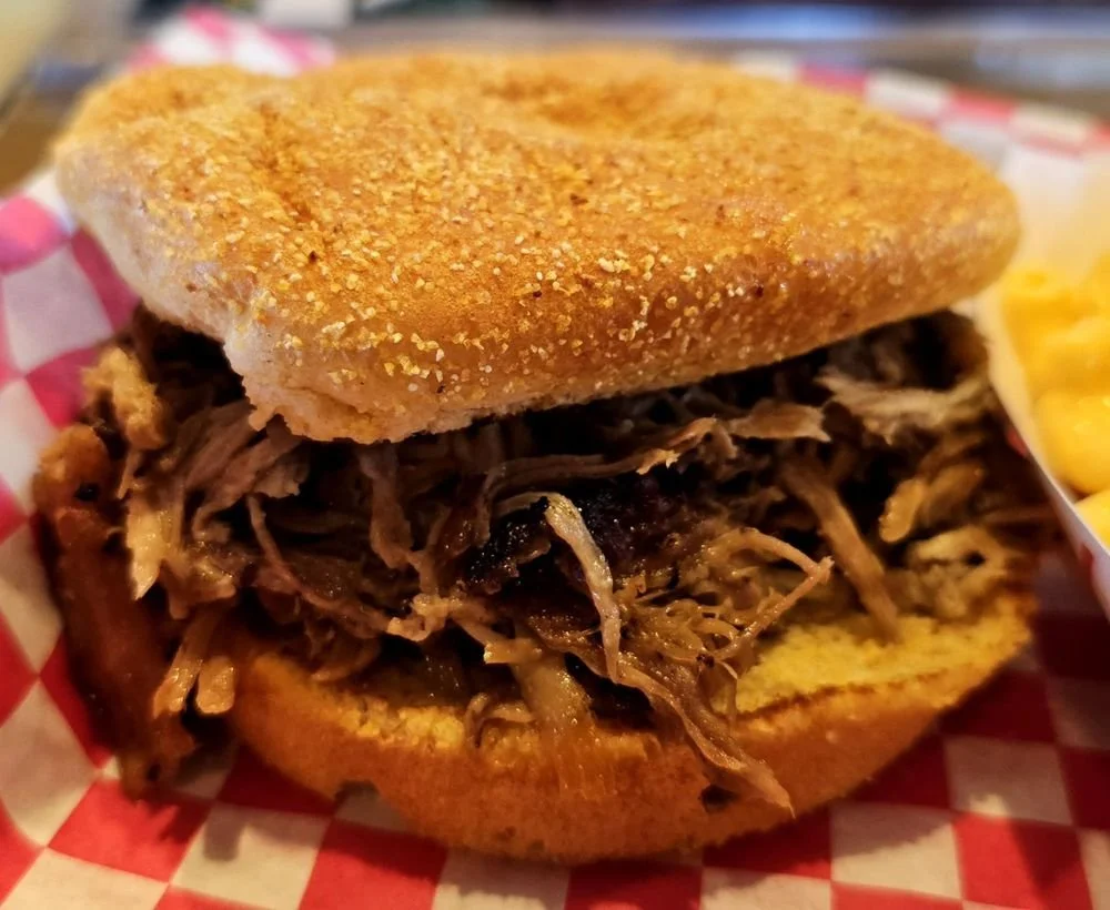 Pulled pork sandwich with bun on a red checkered paper, served with a side of macaroni and cheese.