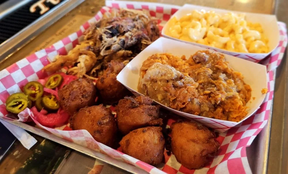 Basket of fried chicken, hush puppies, jalapenos, and pickled red onions, with macaroni and cheese and gravy in small bowls, on a red and white checkered paper-lined tray.