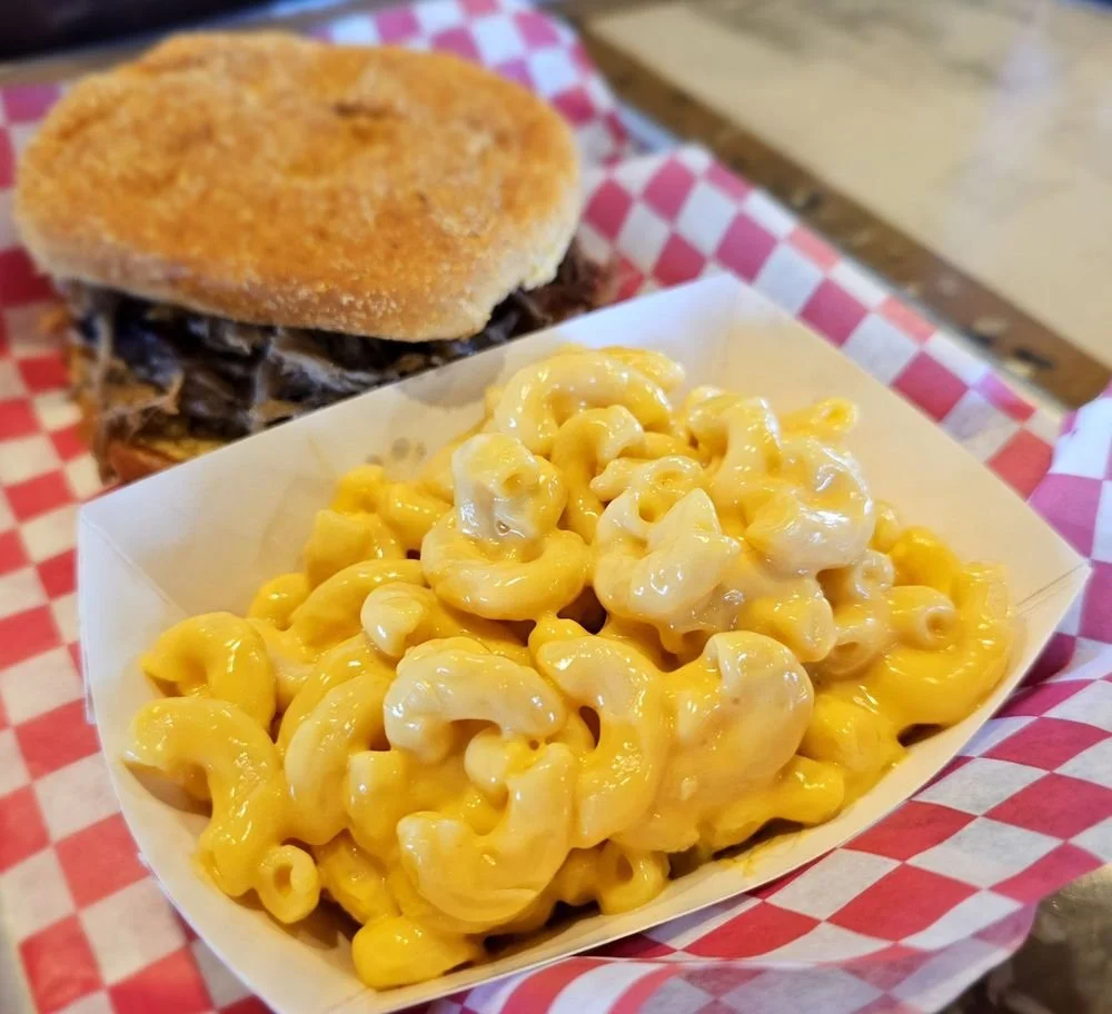 Macaroni and cheese in a white tray with a sandwich in the background on a red and white checkered paper.