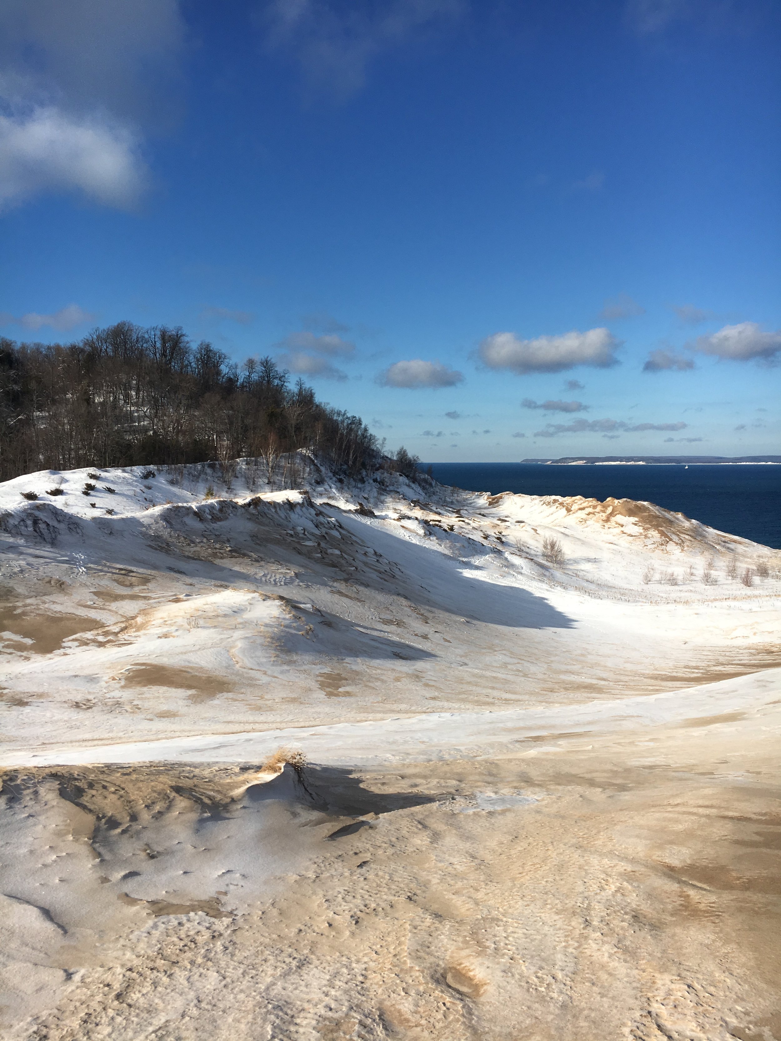 Hilly sand dune landscape blanketed by a light dusting of snow with bare-branched deciduous trees and Lake Michigan in the distance. Intensely blue sky with scattered cloud cover.