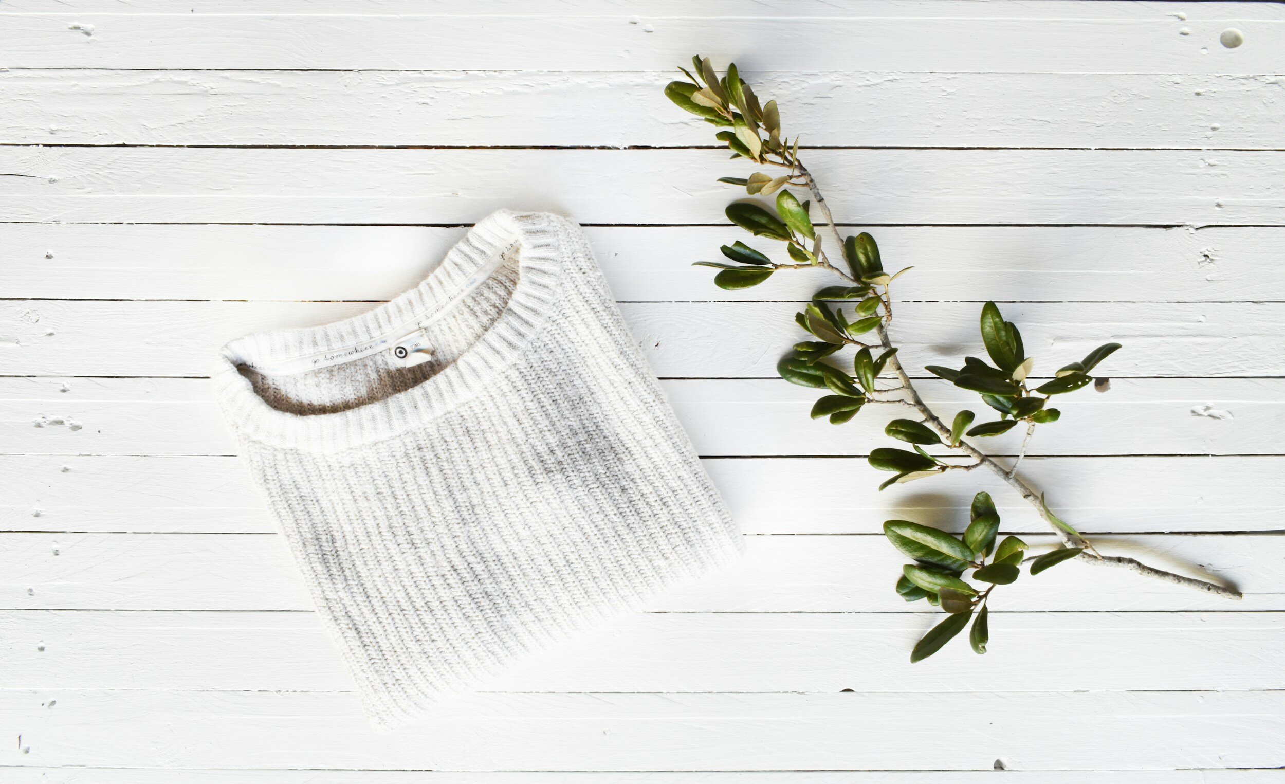 Folded white knitted sweater next to an olive branch on a white wooden surface.
