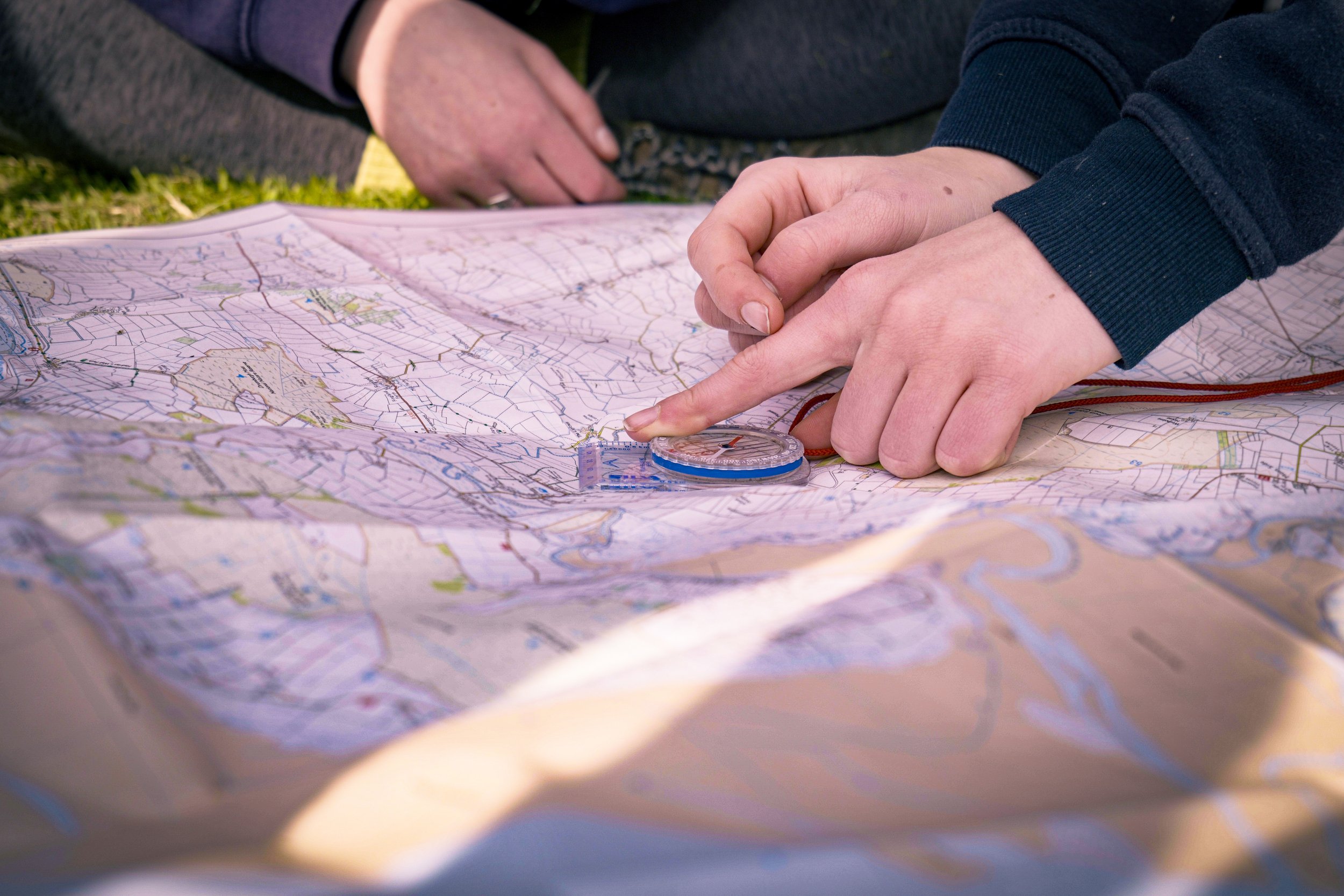 A person using a compass to navigate on a map outdoors, with another person visible in the background.