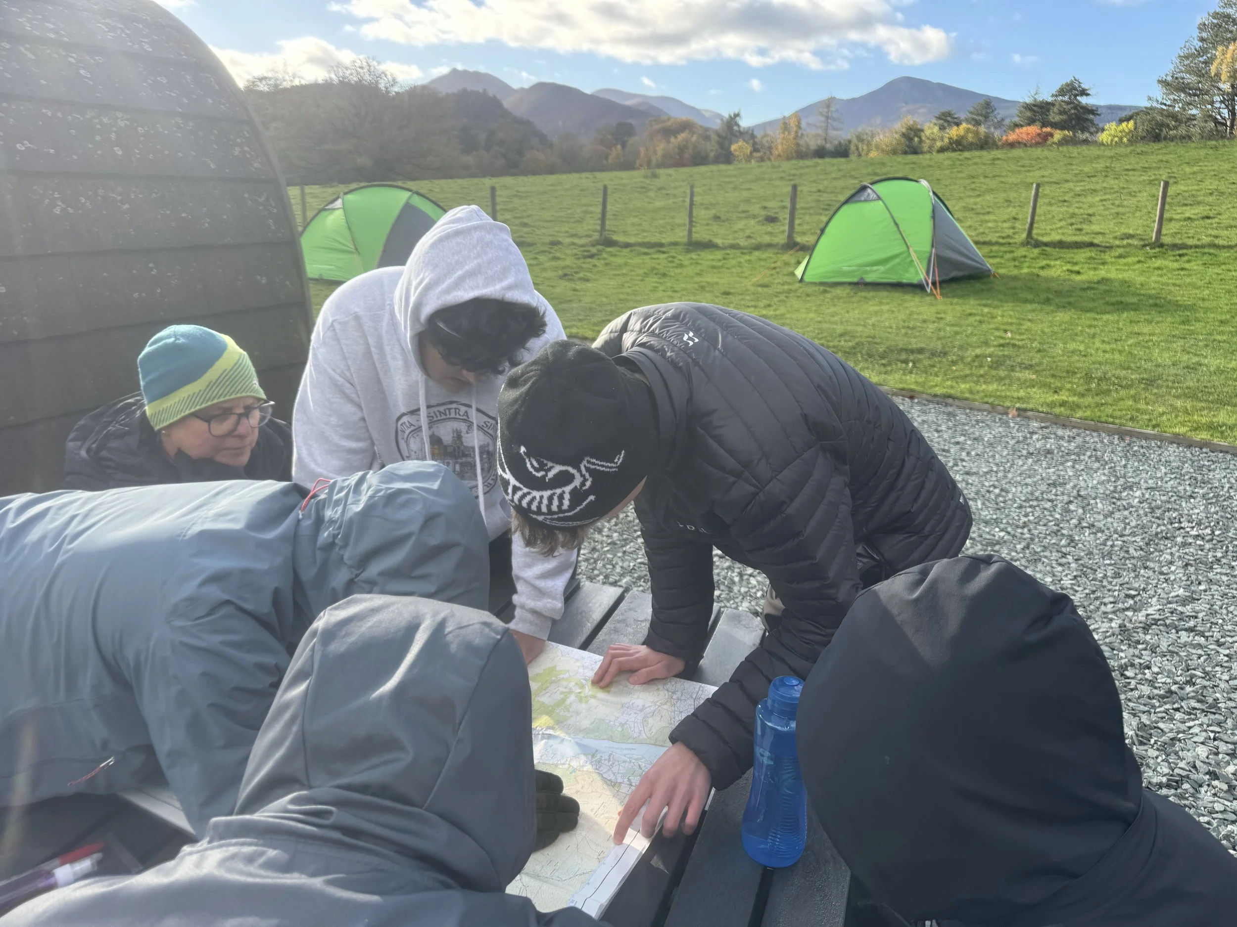 Group of people gathered around a map on a table outdoors, surrounded by tents on a grassy field with mountains in the background.