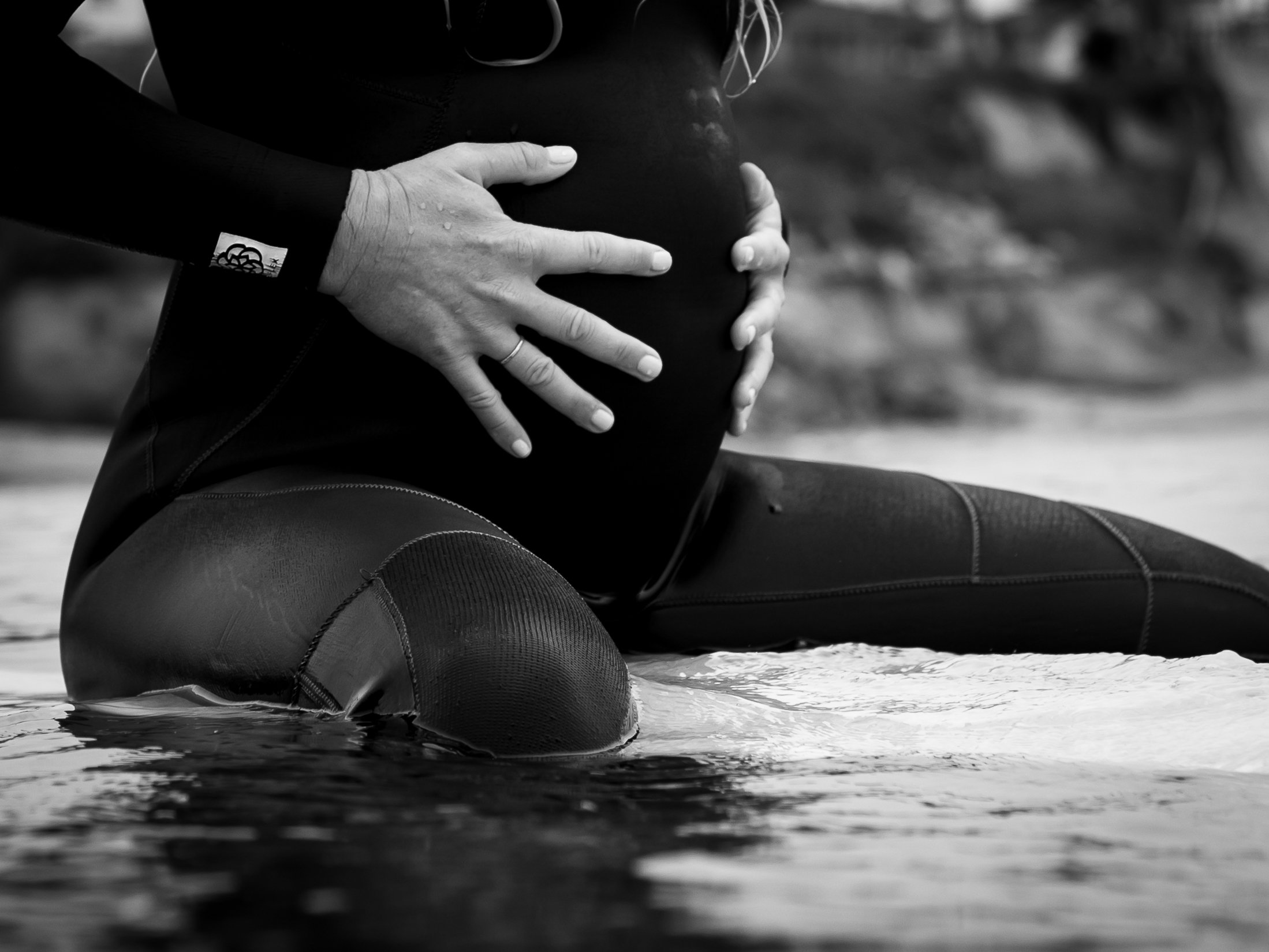 A pregnant person in a wetsuit kneeling on the edge of the water, gently cradling their belly with both hands.