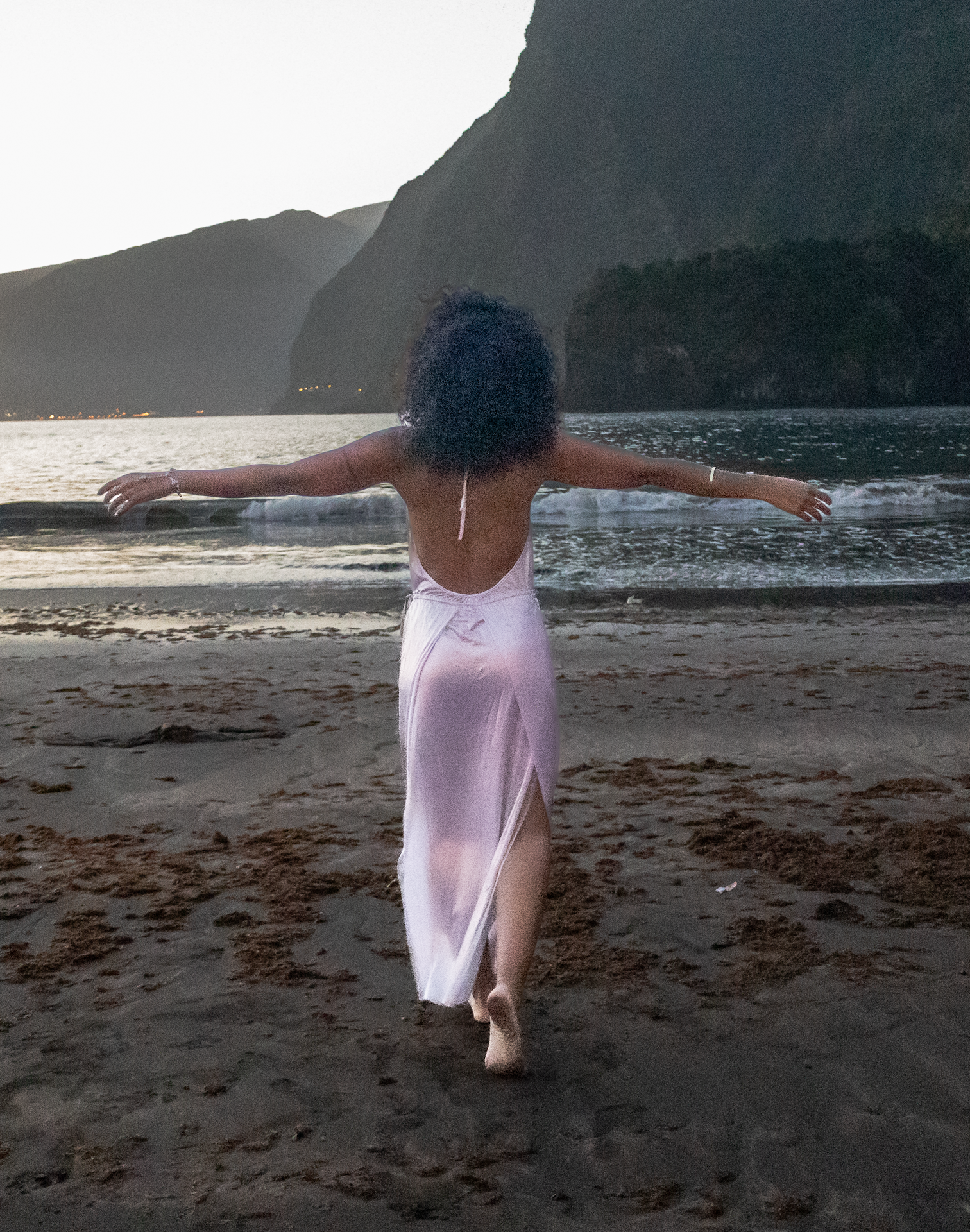Woman in a flowing dress walking barefoot on a beach with arms outstretched at sunset, mountains and ocean in the background.