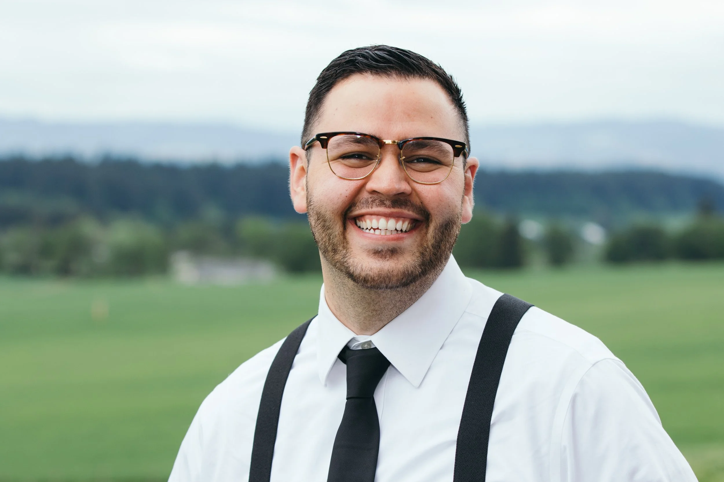 A smiling man with glasses, a white shirt, black suspenders, and a black tie stands outdoors with a grassy field and hills in the background.