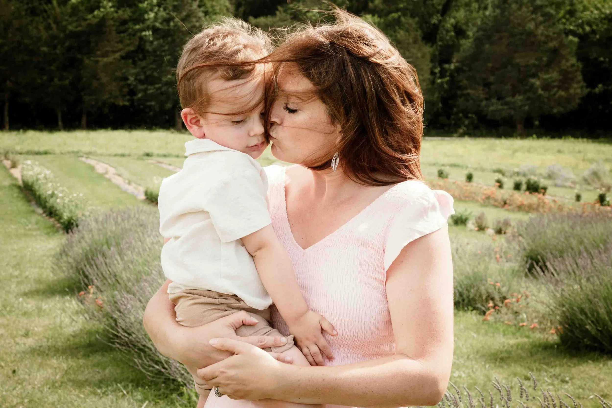 Mother kissing her young son on the cheek during a summer family session at Seven Oaks Lavender Farm in Fauquier County, Virginia with lavender fields in the background.