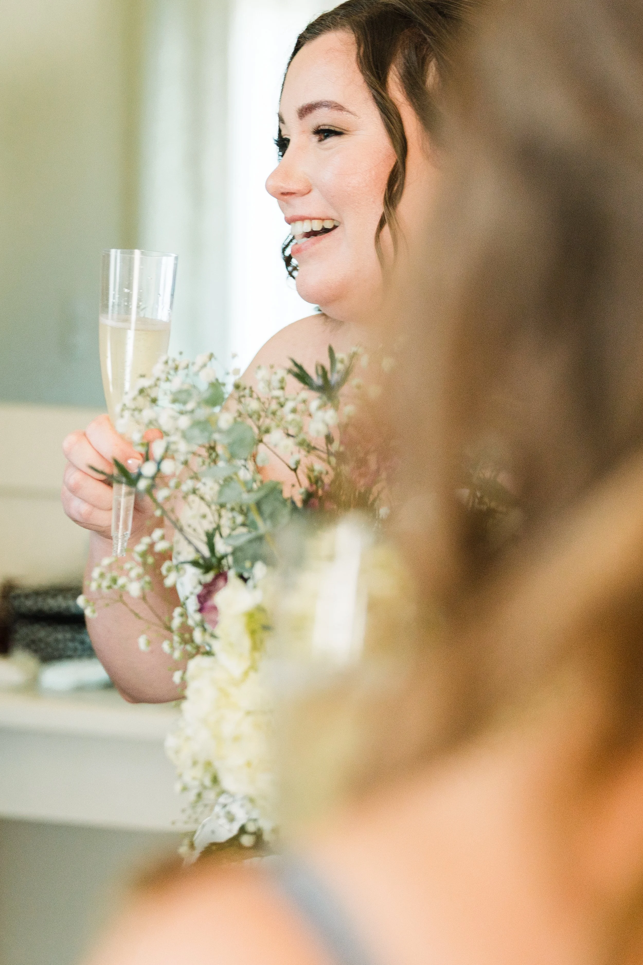 Bride laughing while holding champagne and bouquet during getting ready at Persimmon Creek Barn in Hanover County, Virginia.