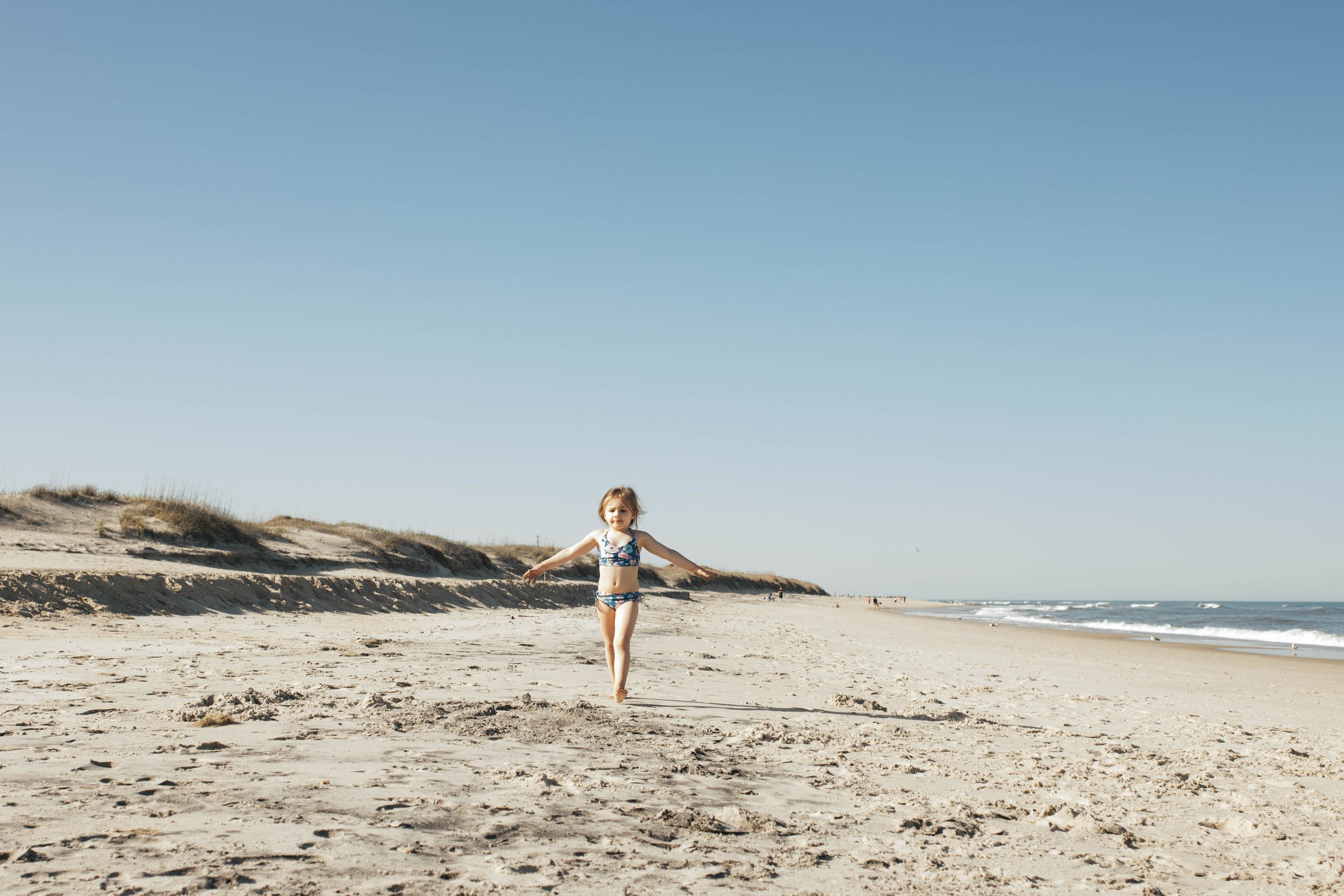 A young girl in a swimsuit running on a sandy beach with the ocean and blue sky in the background.
