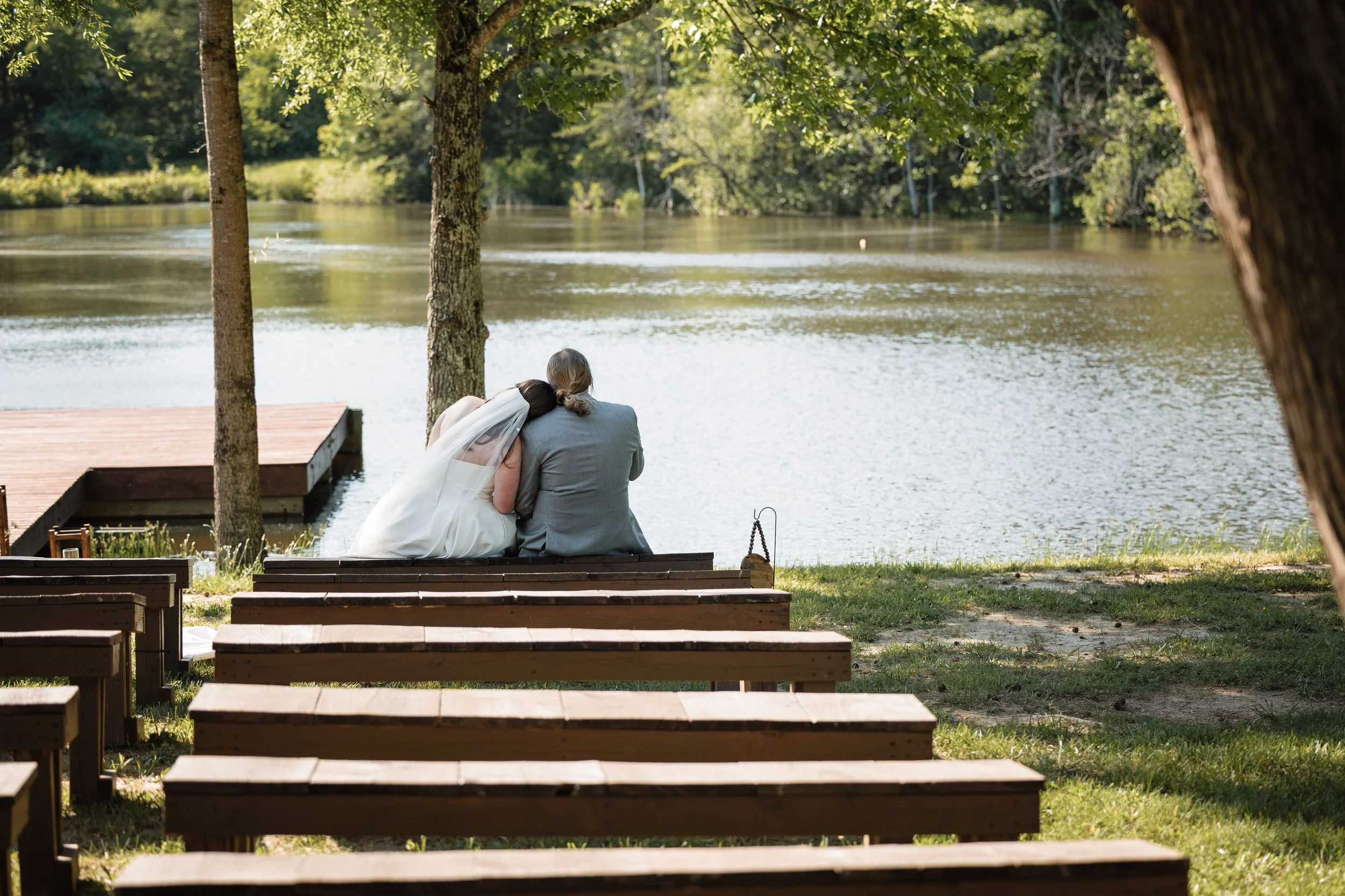 Bride and groom sitting together on wooden ceremony benches by the lake at Persimmon Creek Barn in Virginia after their outdoor wedding ceremony.