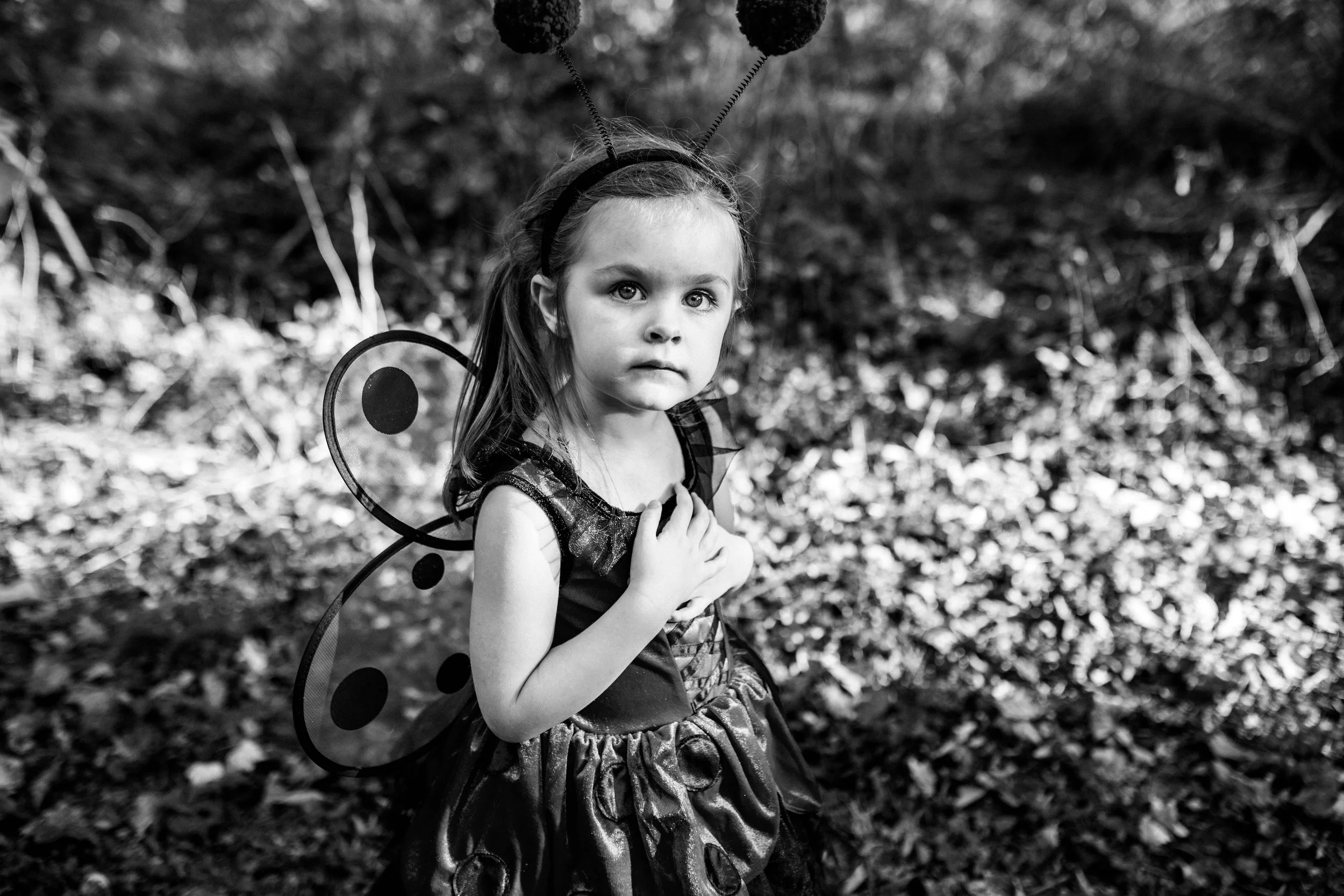 A young girl dressed in a ladybug costume with wings and antennae standing outdoors on a leaf-covered ground, looking at the camera.