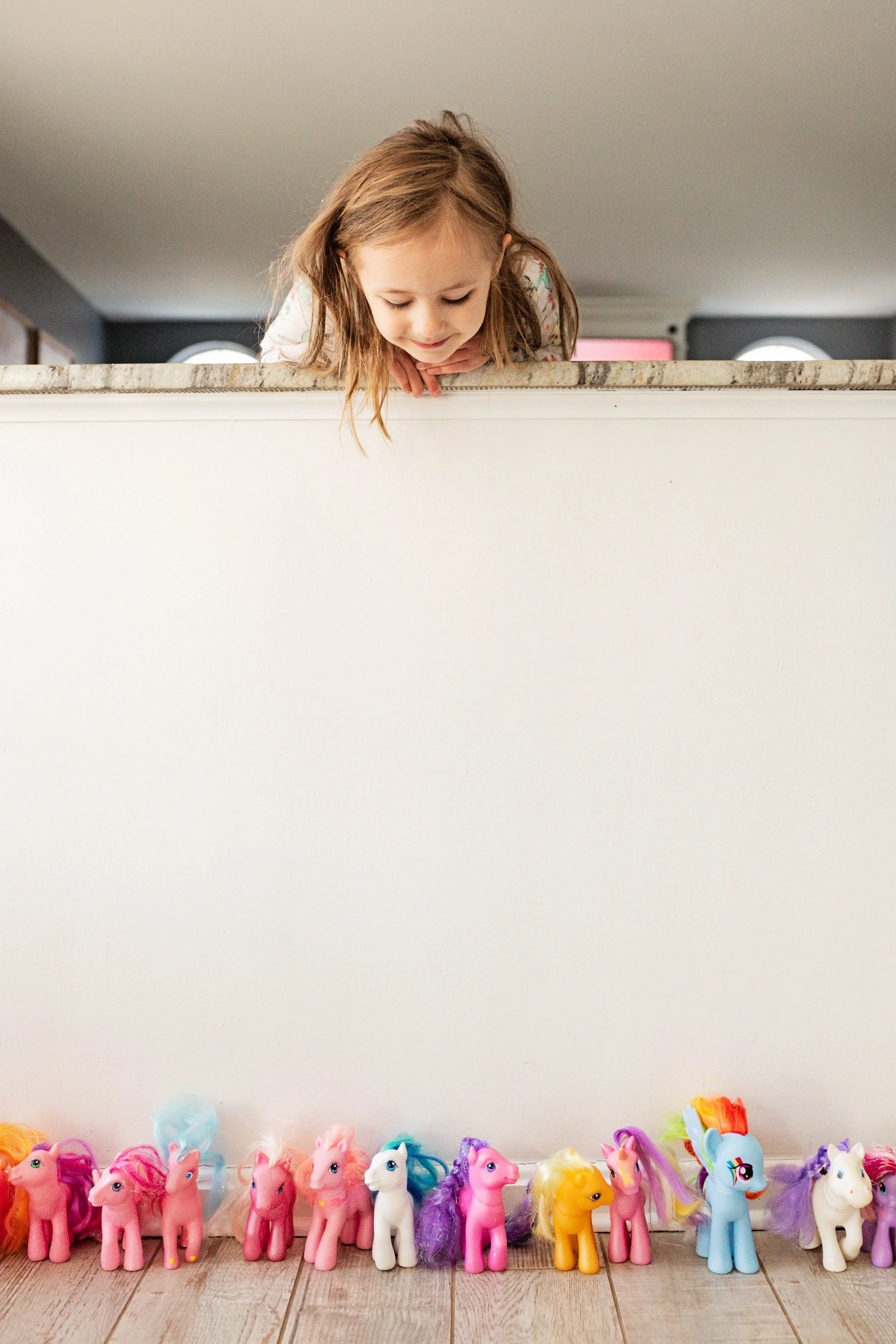 A young girl looking over a white wall at a row of colorful toy horses lined up on a wooden floor.