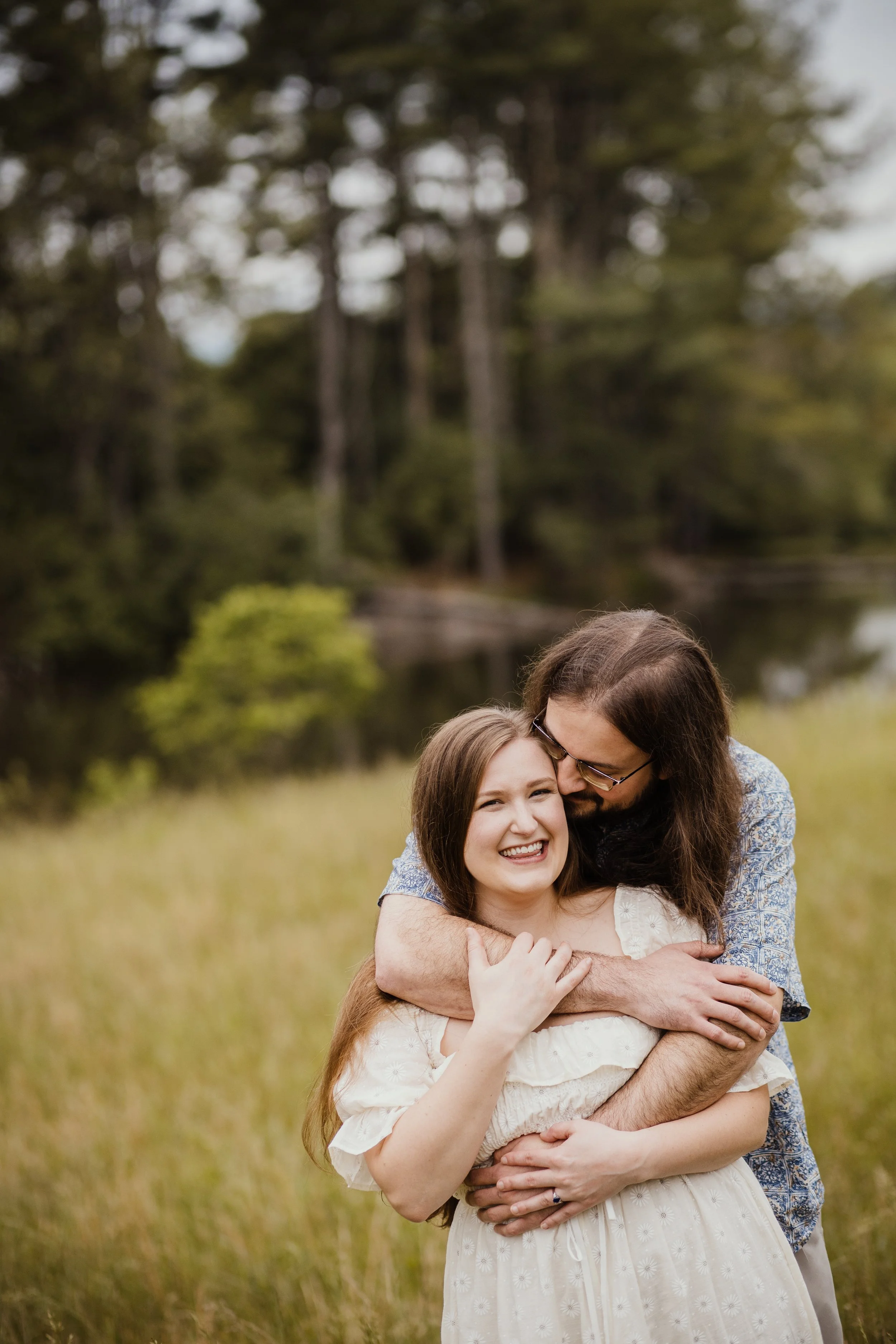 A young woman with long red hair and a white dress smiling as she is embraced by a man with long brown hair and glasses, in an outdoor setting with trees and grass in the background.