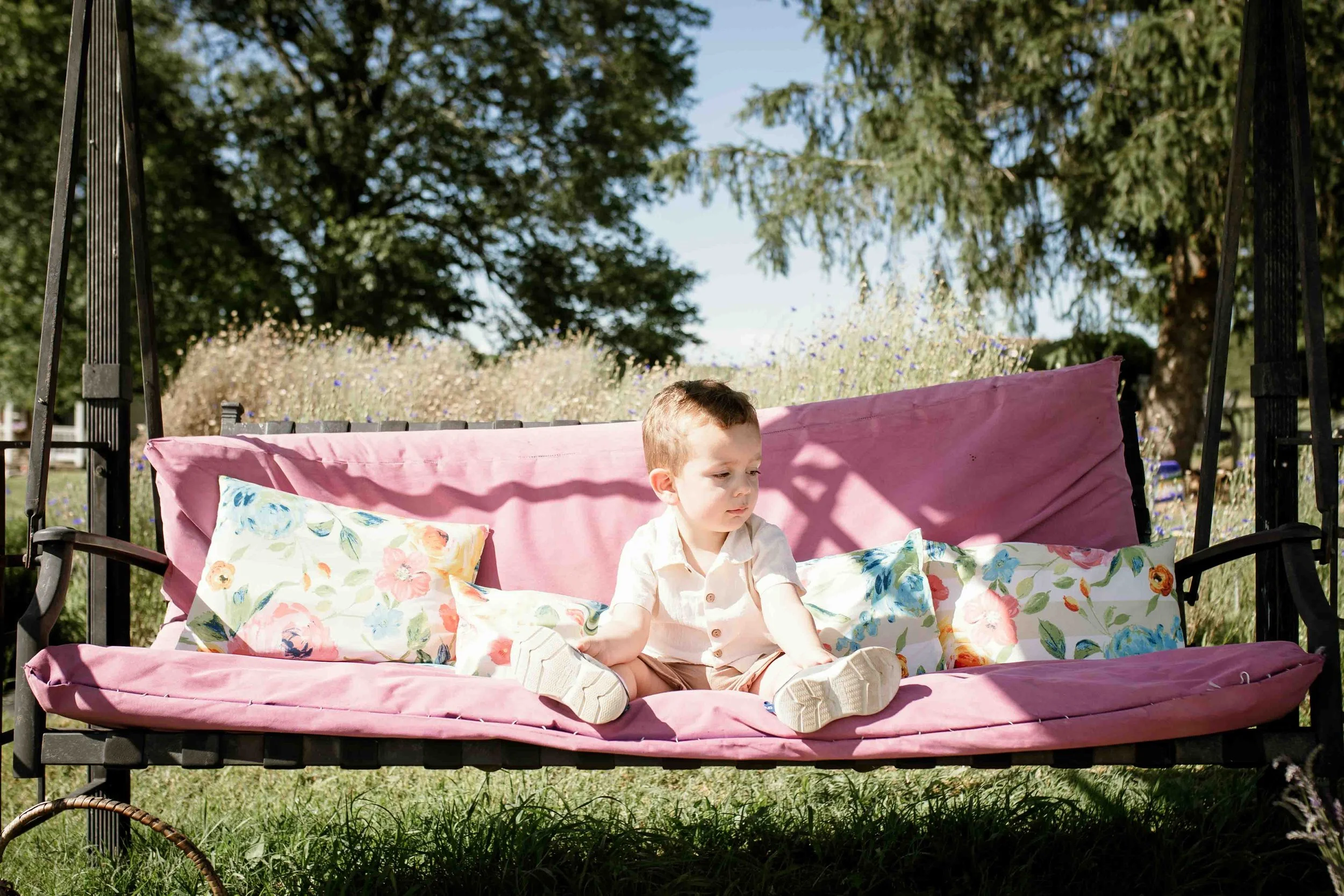 Toddler boy sitting on a pink vintage swing with floral pillows at Seven Oaks Lavender Farm in Fauquier County, Virginia during a summer family session.