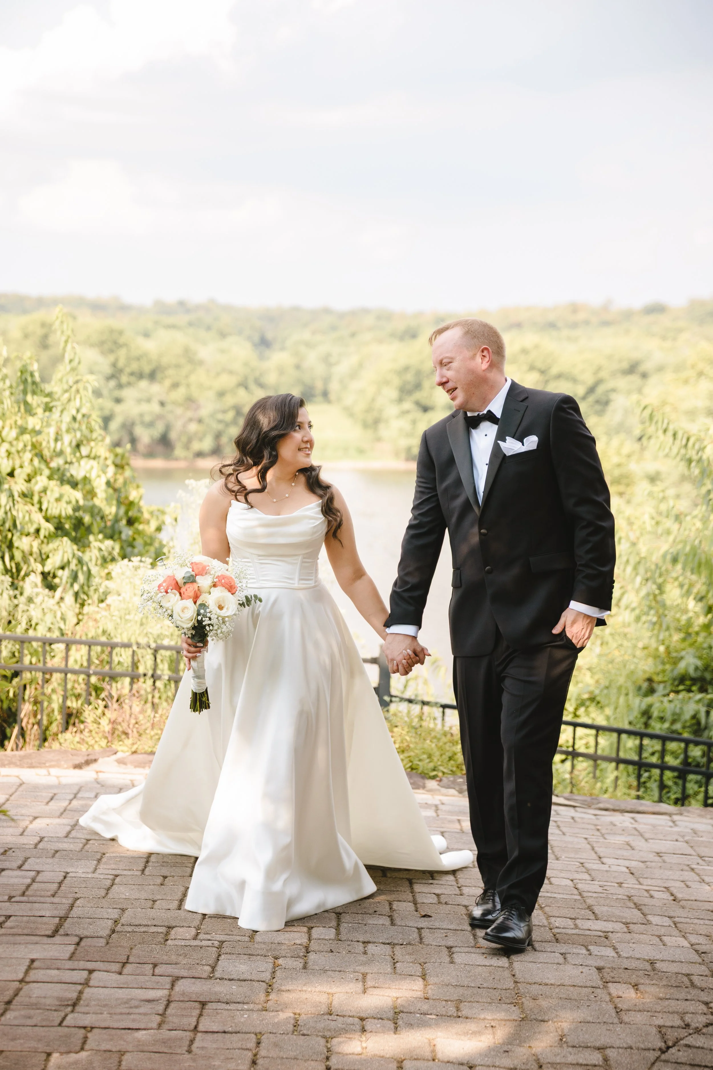 A bride and groom walking hand in hand outdoors near a lake, surrounded by lush green trees, on their wedding day.