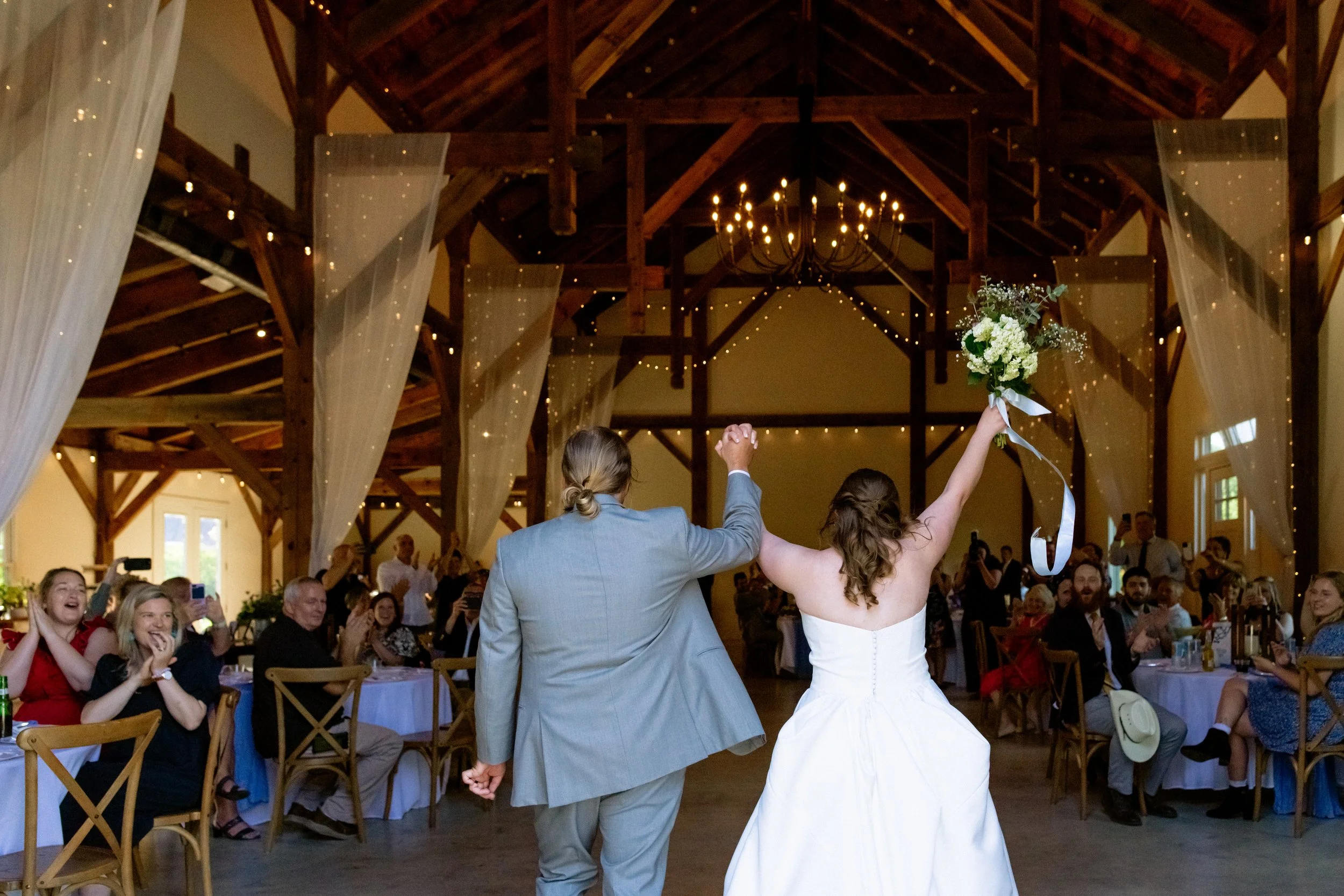 Bride and groom entering their reception at Persimmon Creek Barn in Virginia with guests cheering under wooden beams and warm string lights.