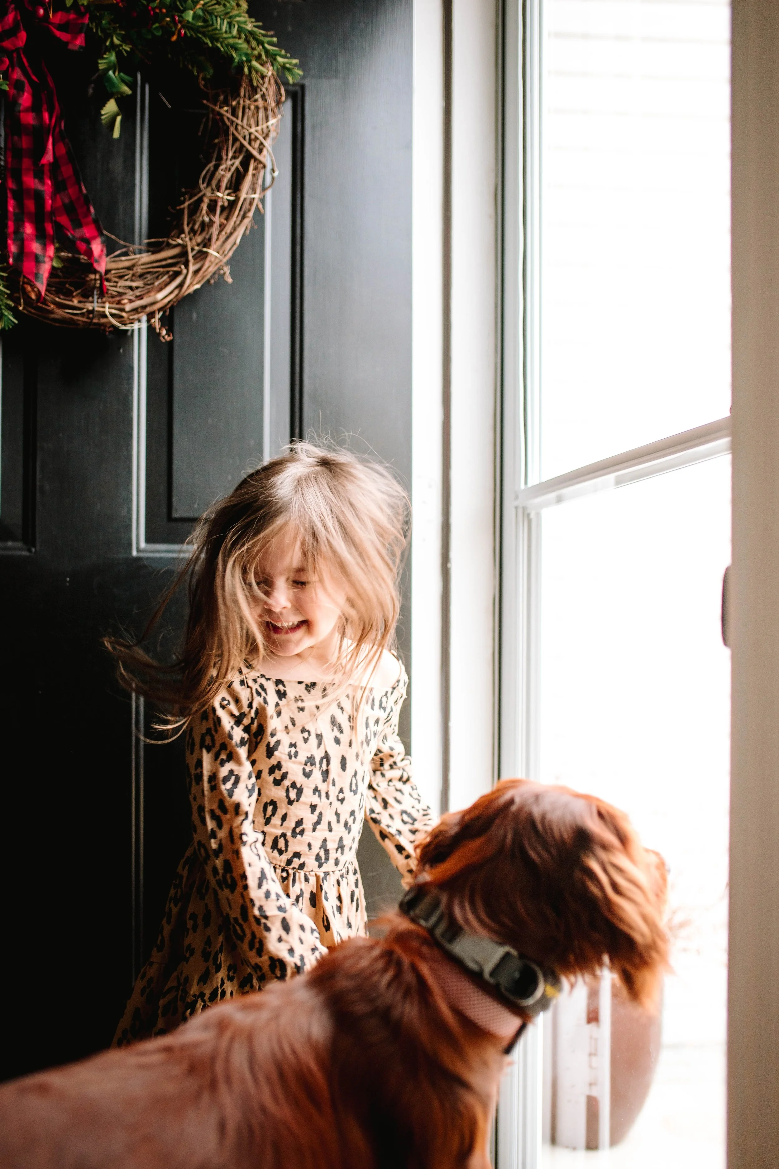 A young girl with long, tousled hair wearing a leopard print dress, smiling and playing with a brown dog by a door with a window, decorated with a festive wreath.