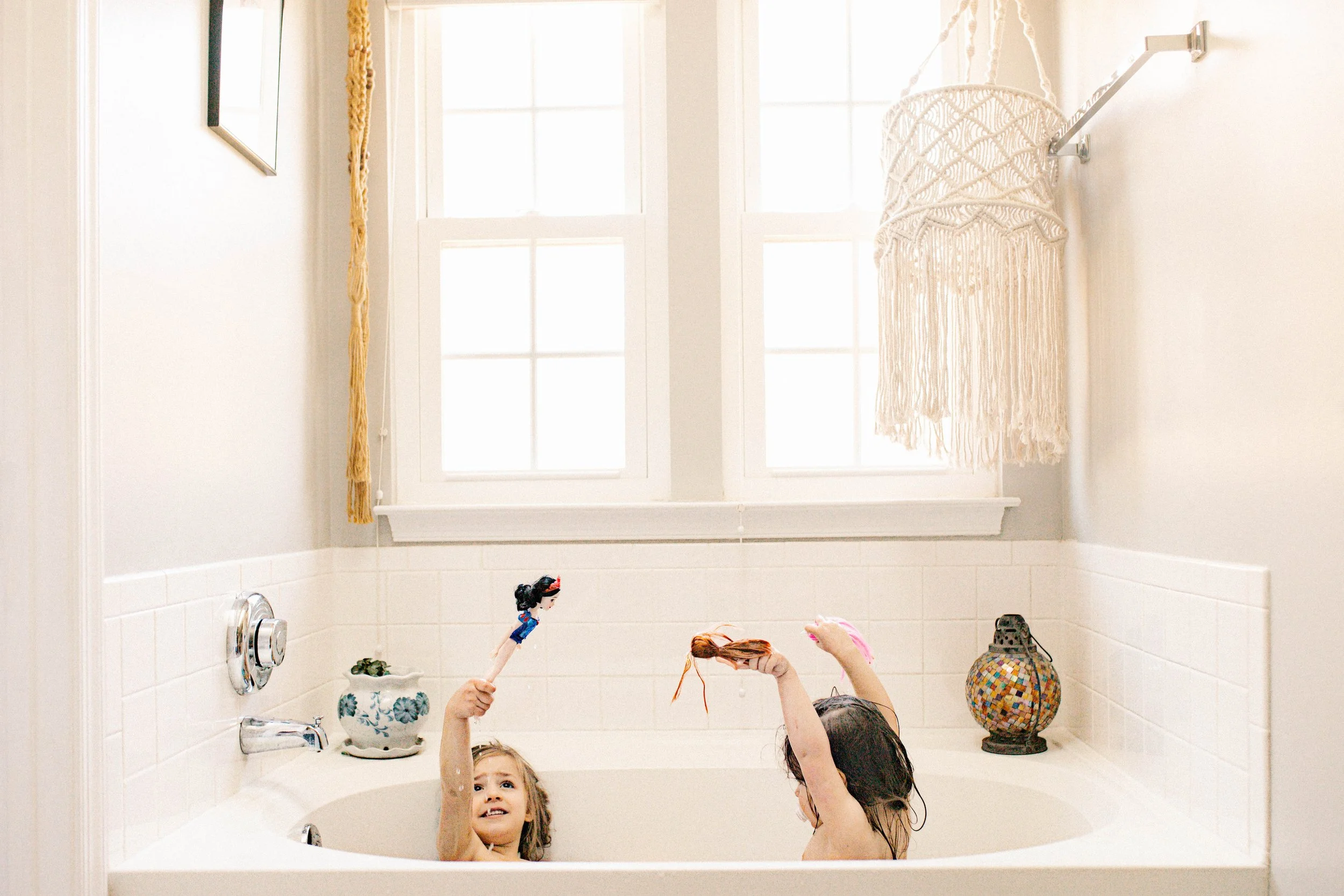 Two children playing in a bathtub filled with water in a bathroom with white walls, a window, and decorative items.