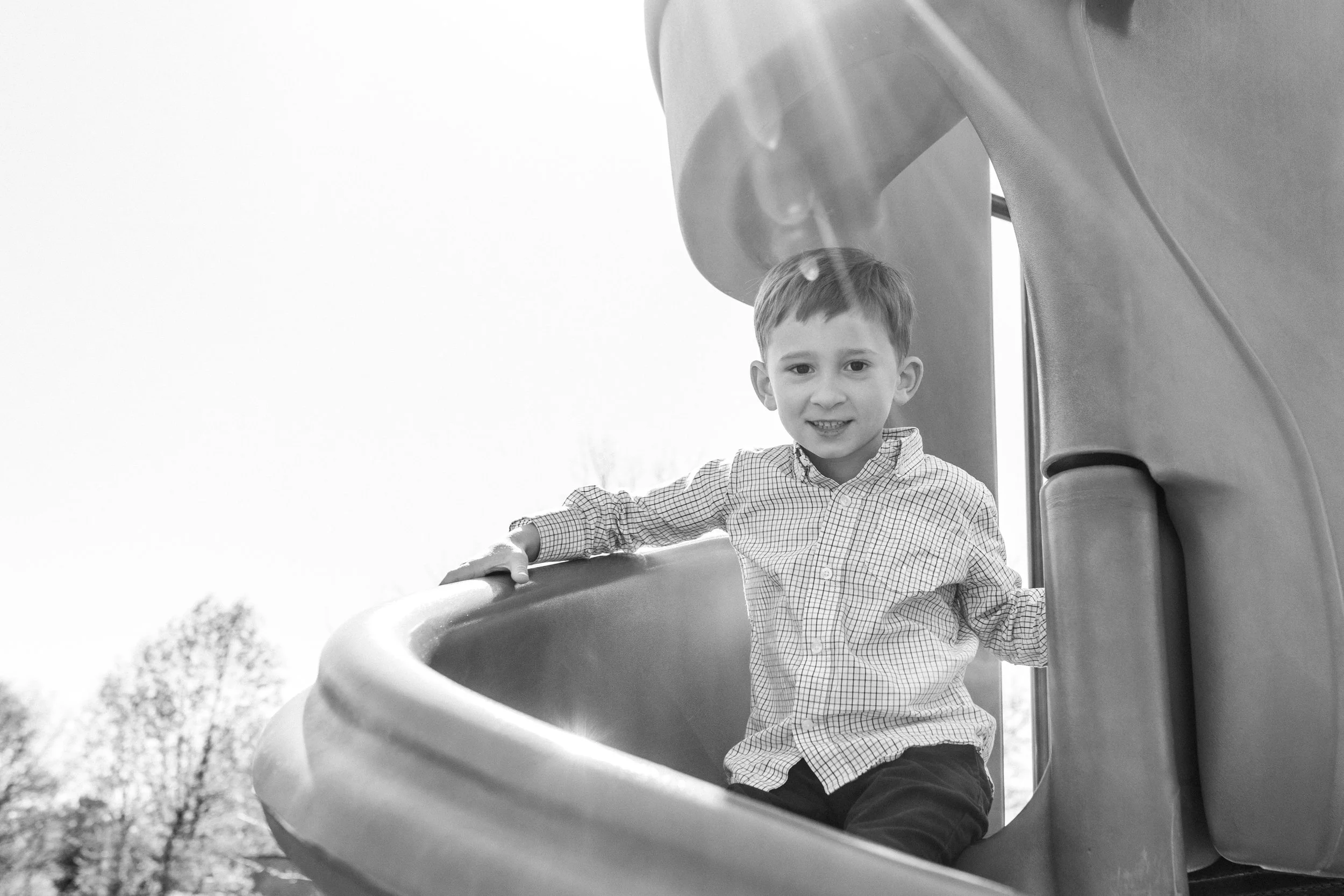 Black and white photo of a young boy smiling at the top of a playground slide during a family session at Rady Park in Fauquier County.