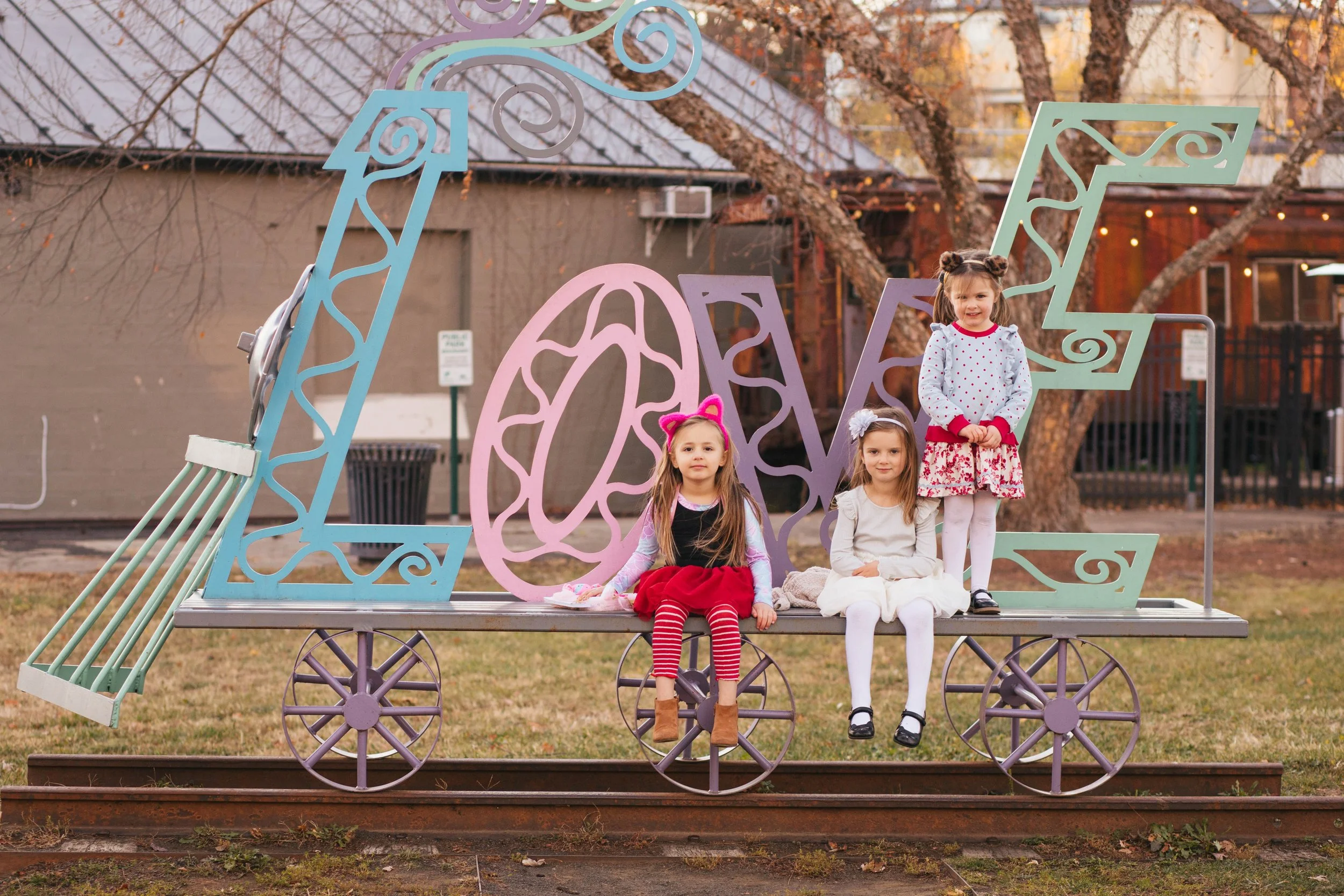 Four young girls sitting on a decorated train car structure outdoors, spelling out the number '2023' in large pastel-colored numbers, with trees and buildings in the background.