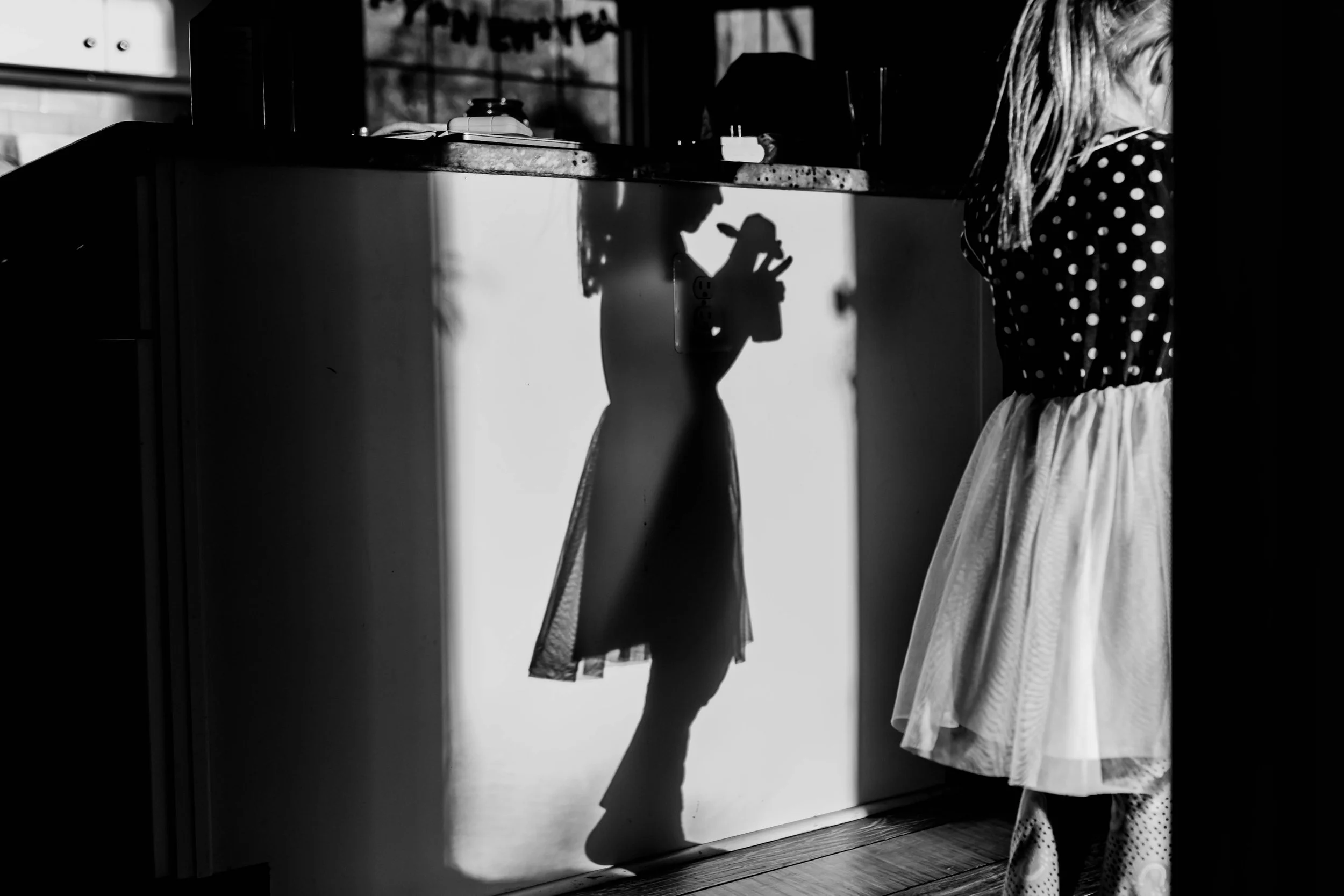 A young girl in a polka-dot top and a white skirt stands near a reflective surface, casting a shadow that resembles her playing with a toy or object, with sunlight creating contrasting shadows in a cozy indoor setting.