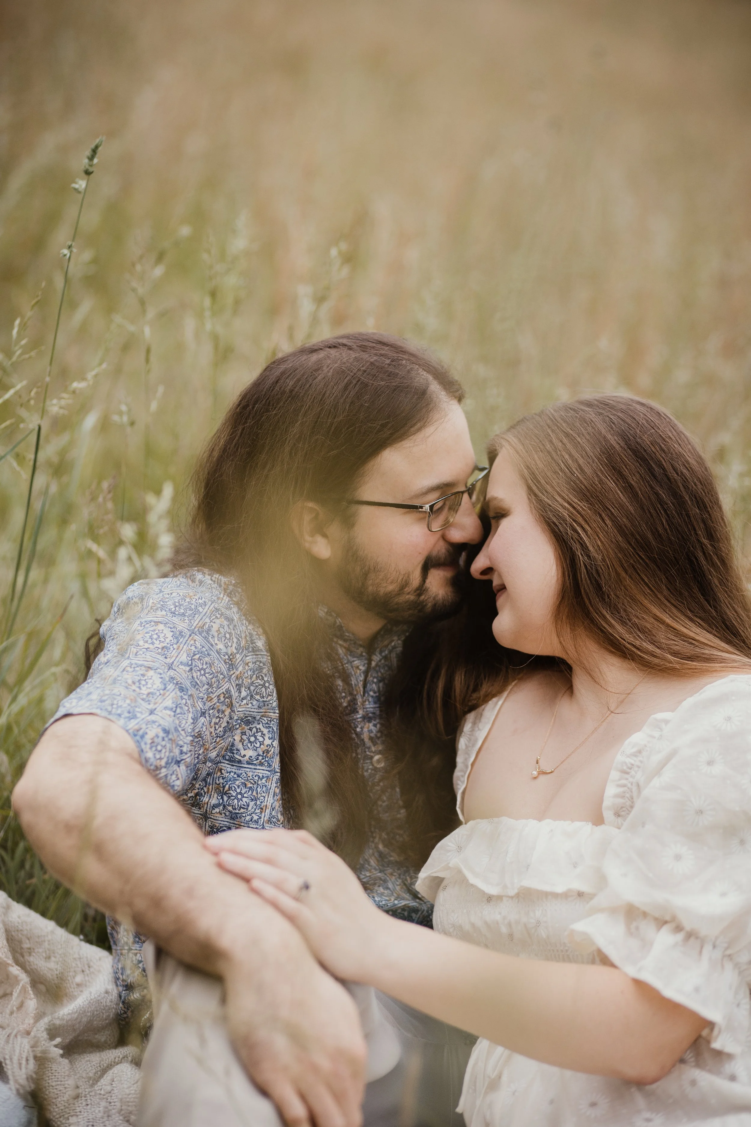 A couple sitting closely in a field of tall, golden grass, touching foreheads with eyes closed, sharing an intimate moment.