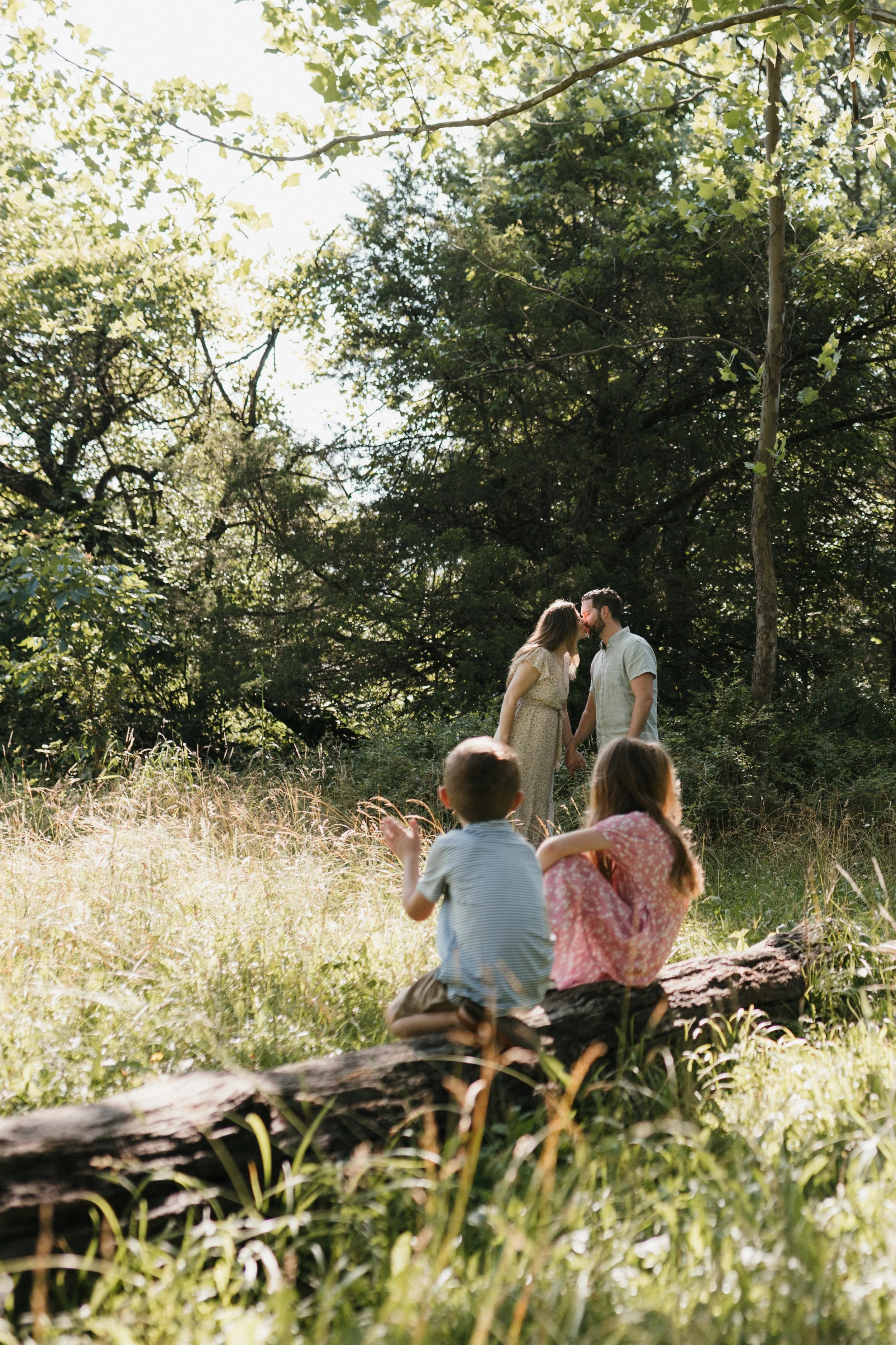 Children sitting on a fallen log watching their parents share a quiet moment during a family photography session at Manassas National Battlefield Park.