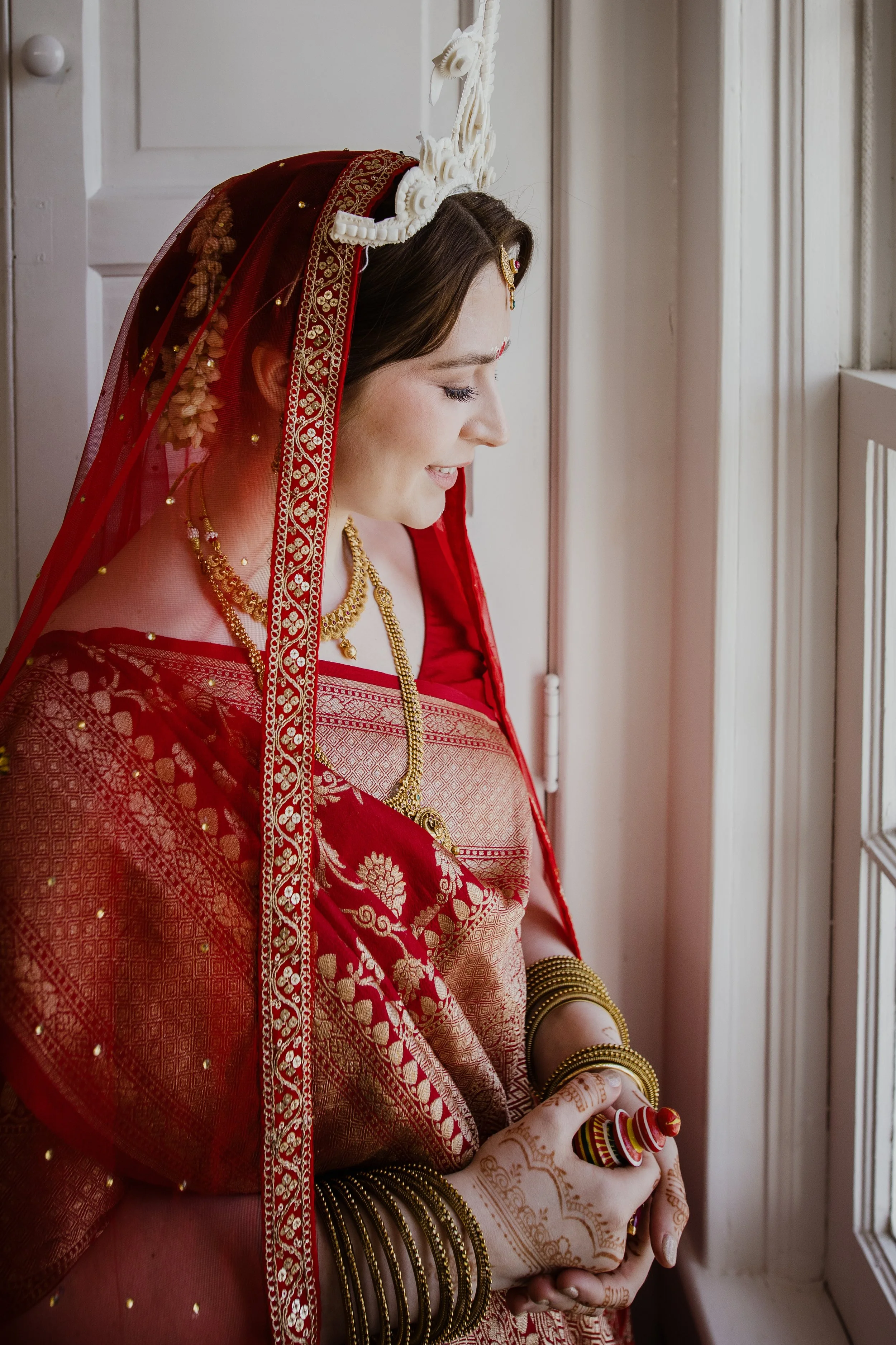 Bride in a red sari and traditional headpiece standing by a window during wedding morning preparations in Northern Virginia.