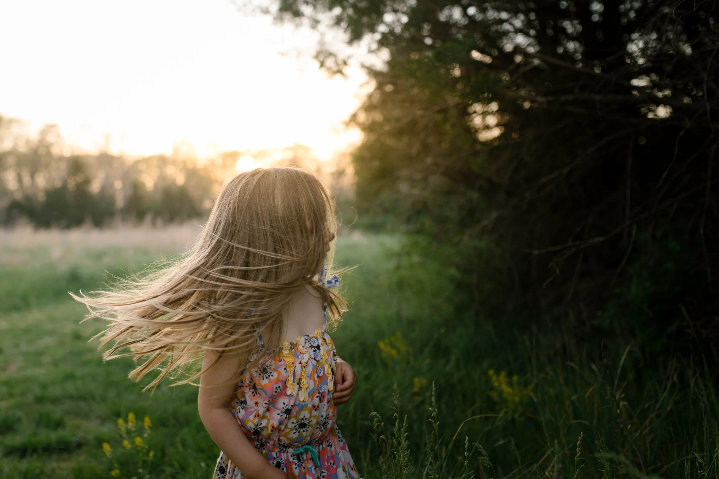 A young girl with long blonde hair standing outdoors in a grassy field at sunset, wearing a colorful dress with a floral pattern.