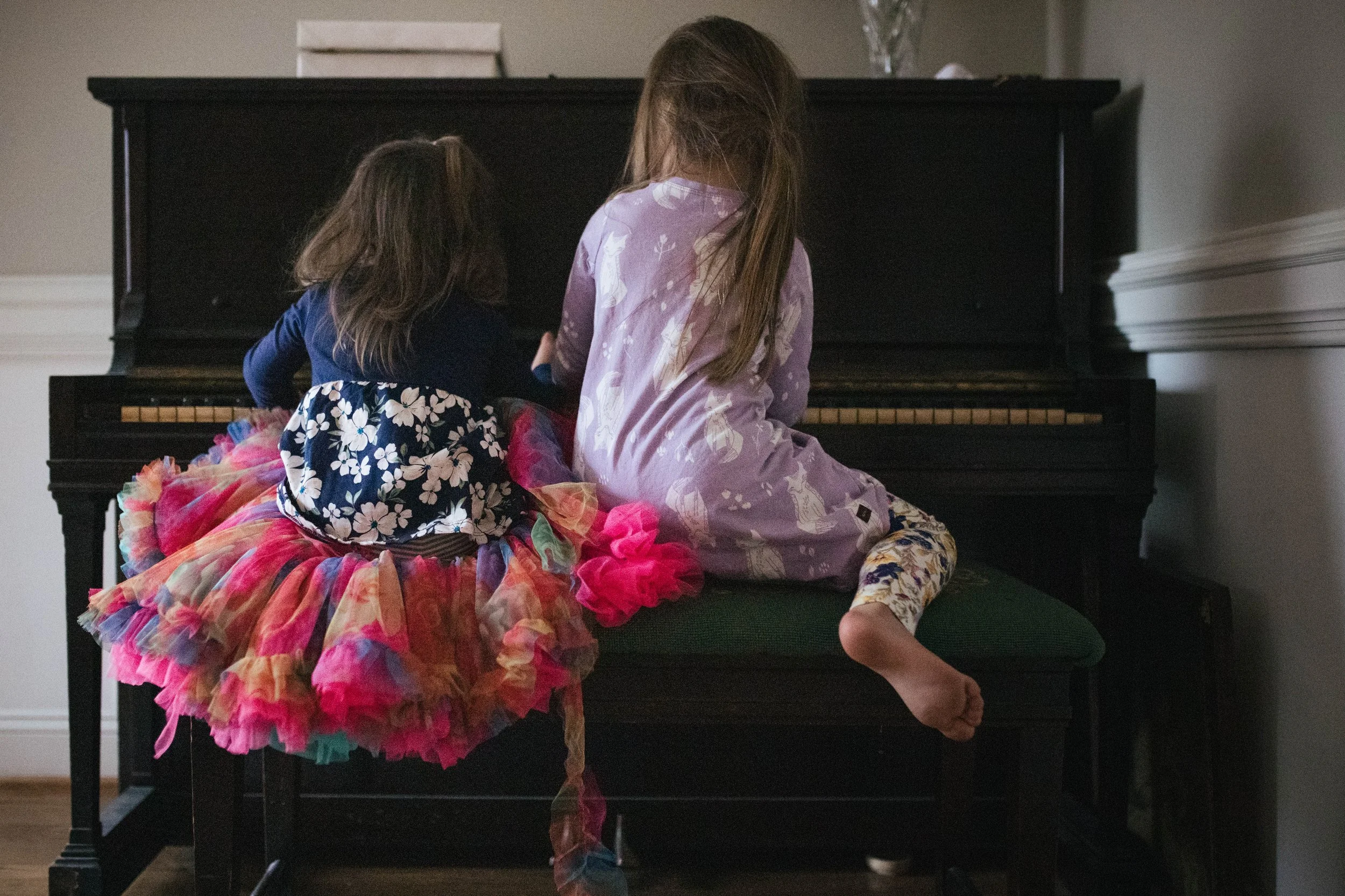 Two young girls, one wearing a colorful tutu and the other in pajamas, sitting on a bench in front of a black upright piano, playing or enjoying time together.