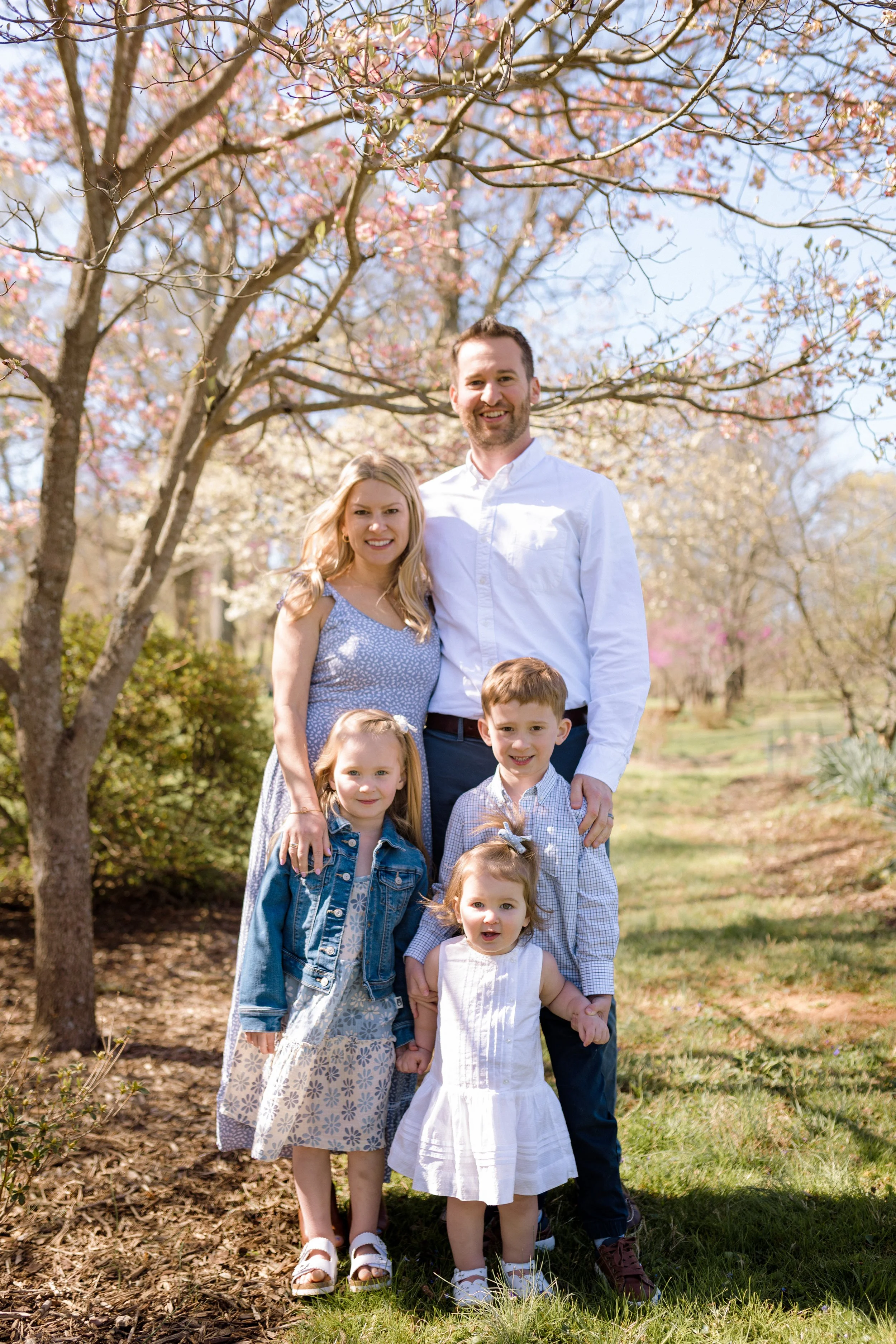 Family of five posing together under blooming trees during a spring morning family photo session at Rady Park in Fauquier County.