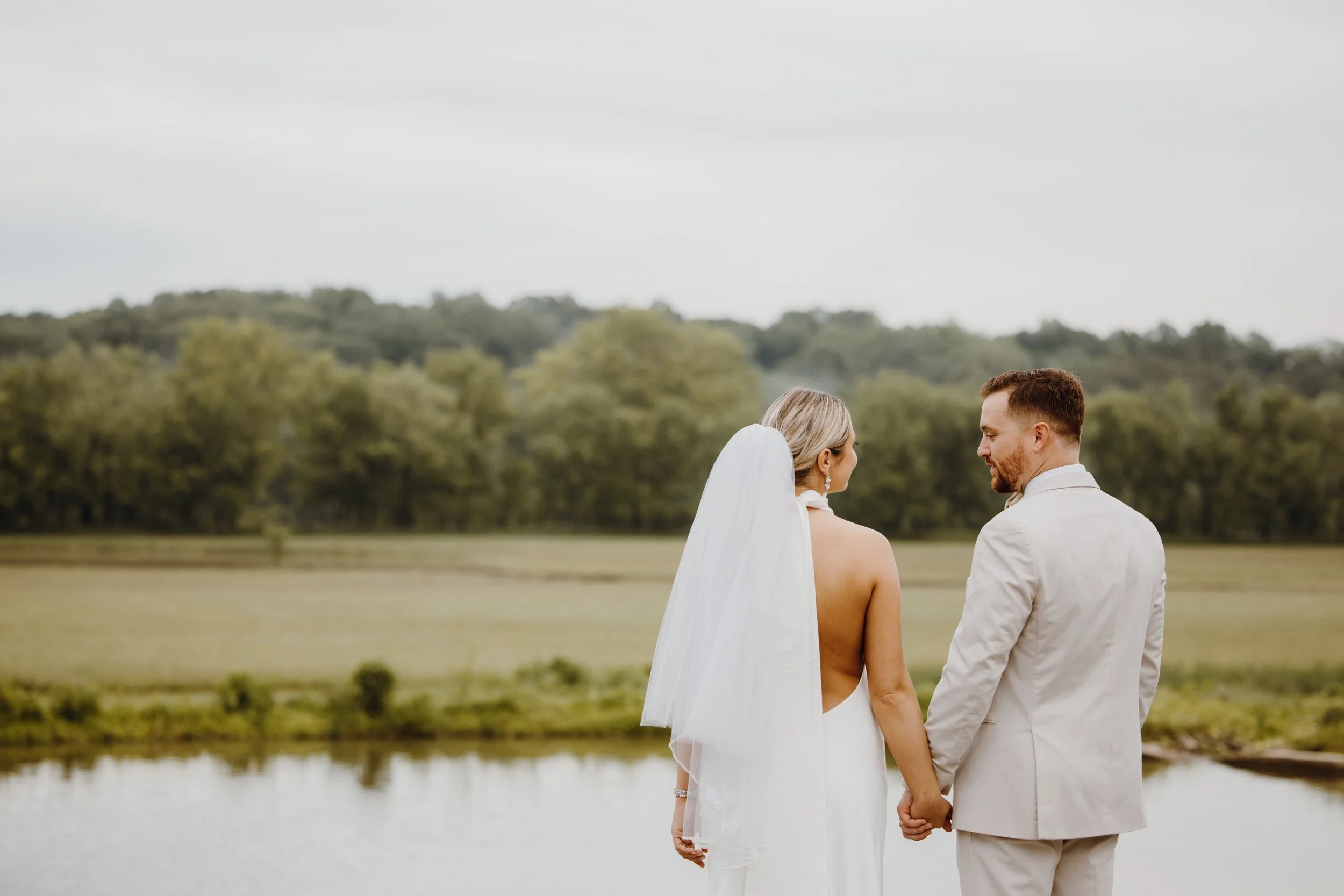 A bride and groom standing hand in hand outdoors near a lake, with a backdrop of trees and a cloudy sky.