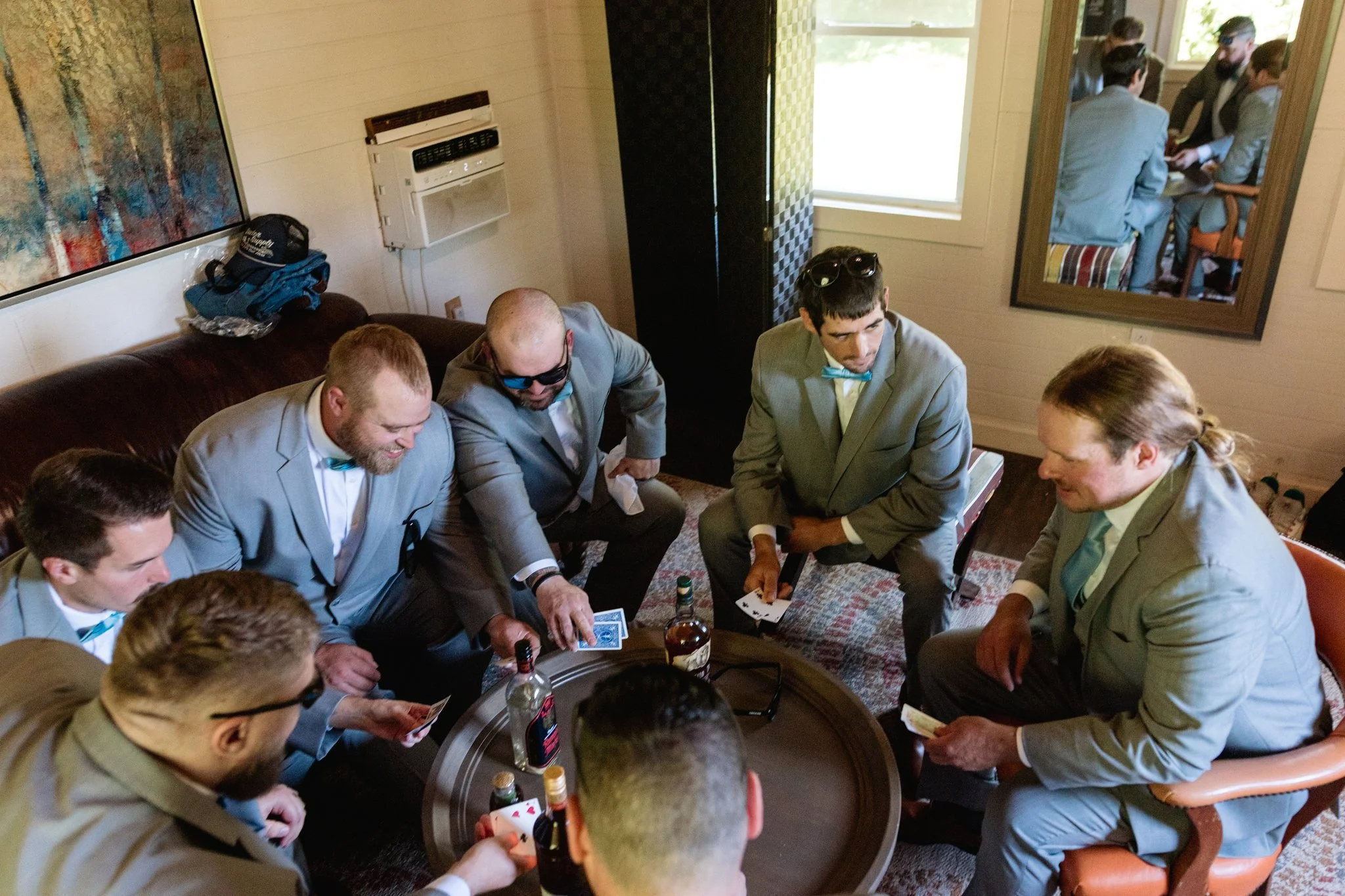 Men in grey suits playing cards around a small table in a room with a patterned carpet, bottles, and a mirror reflecting more men in suits.