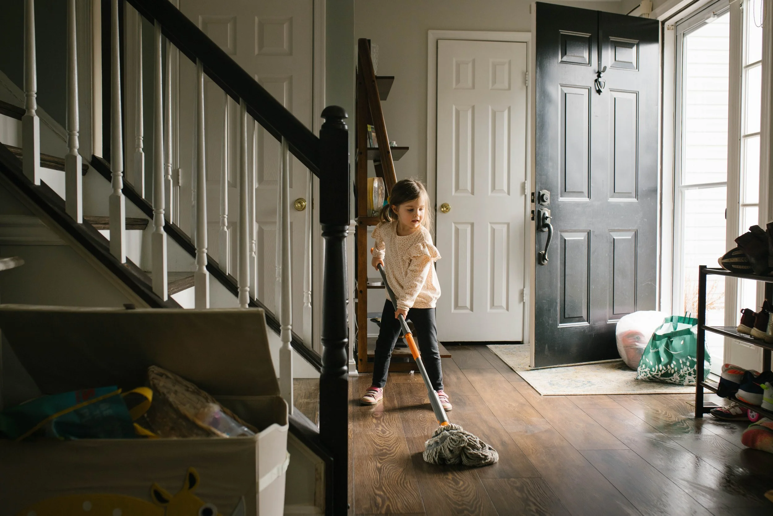 A young girl is vacuuming the wooden floor of a home's entryway near the front door.