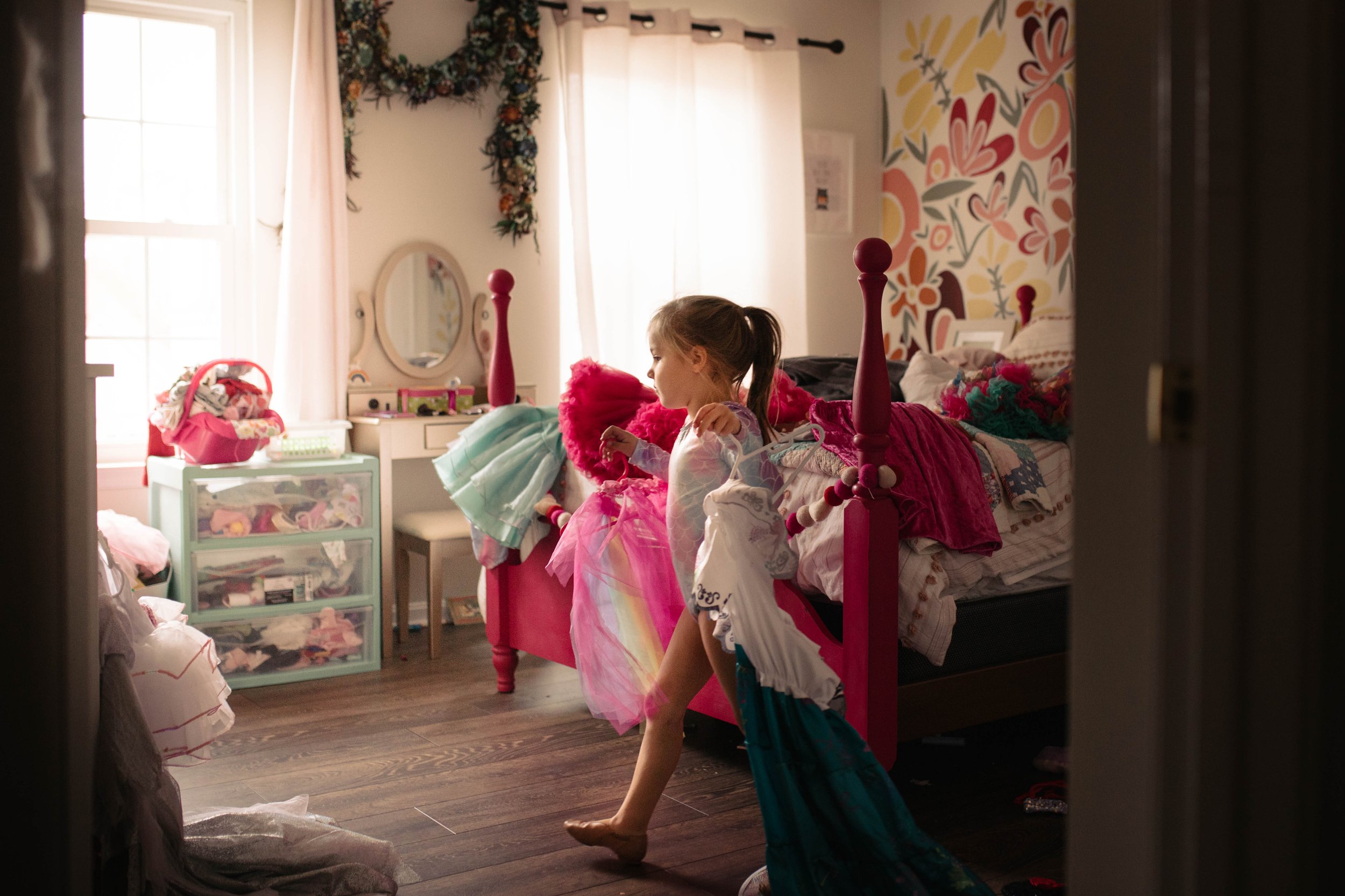 A young girl in pajamas standing on one leg, holding a dress, in a bedroom filled with pink and pastel decorations, toys, and a bed with colorful bedding.