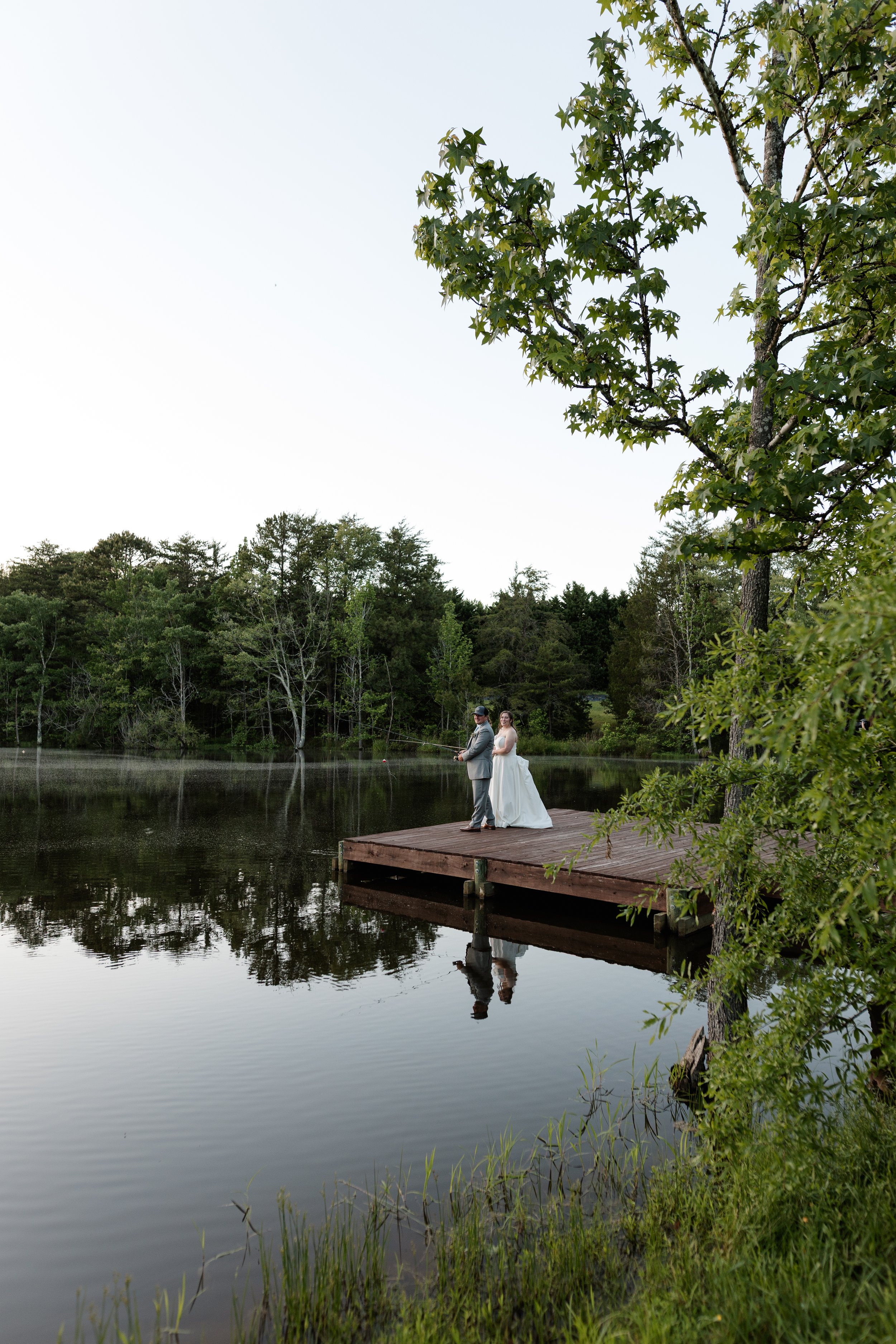 A bride and groom standing on a wooden dock by a lake, fishing at sunset surrounded by trees.
