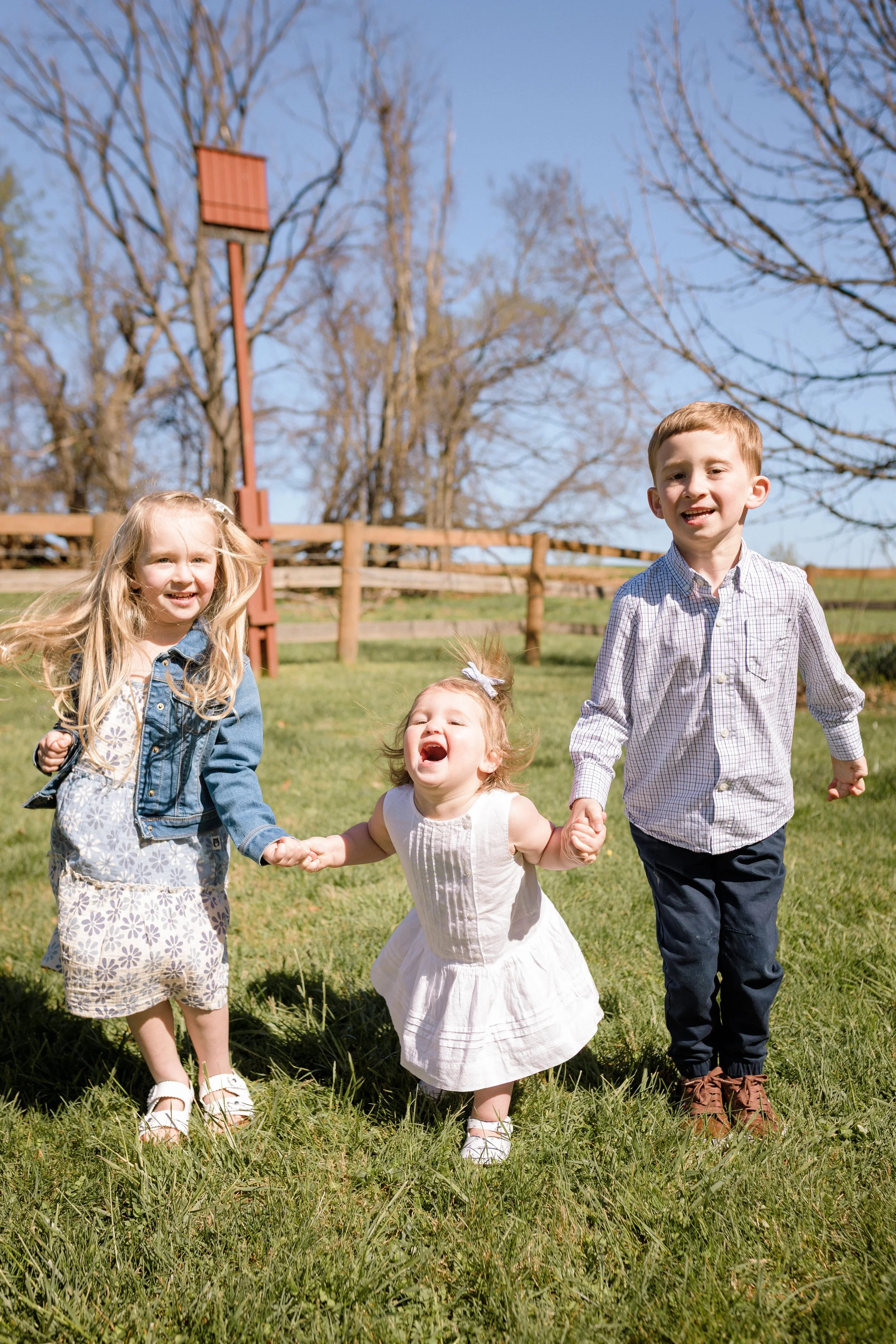 Three young siblings holding hands and laughing while running through the grass during a spring family photo session at Rady Park in Fauquier County.