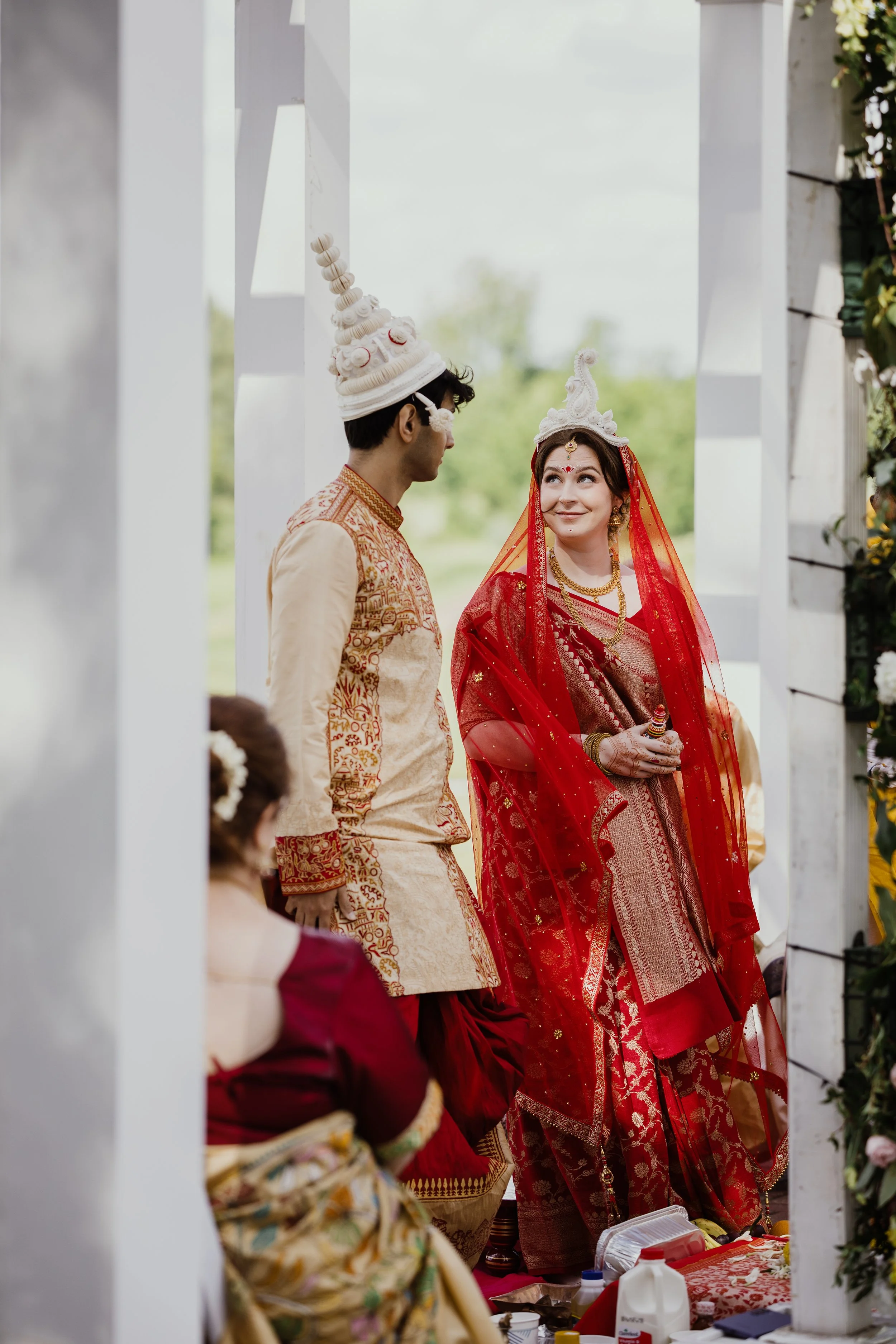 A bride and groom in traditional Indian wedding attire standing under a white canopy with greenery in the background. The bride is wearing a red saree with gold embroidery and a white ceremonial headpiece, and the groom is wearing beige and gold embroidered traditional clothing with a tall, white turban.