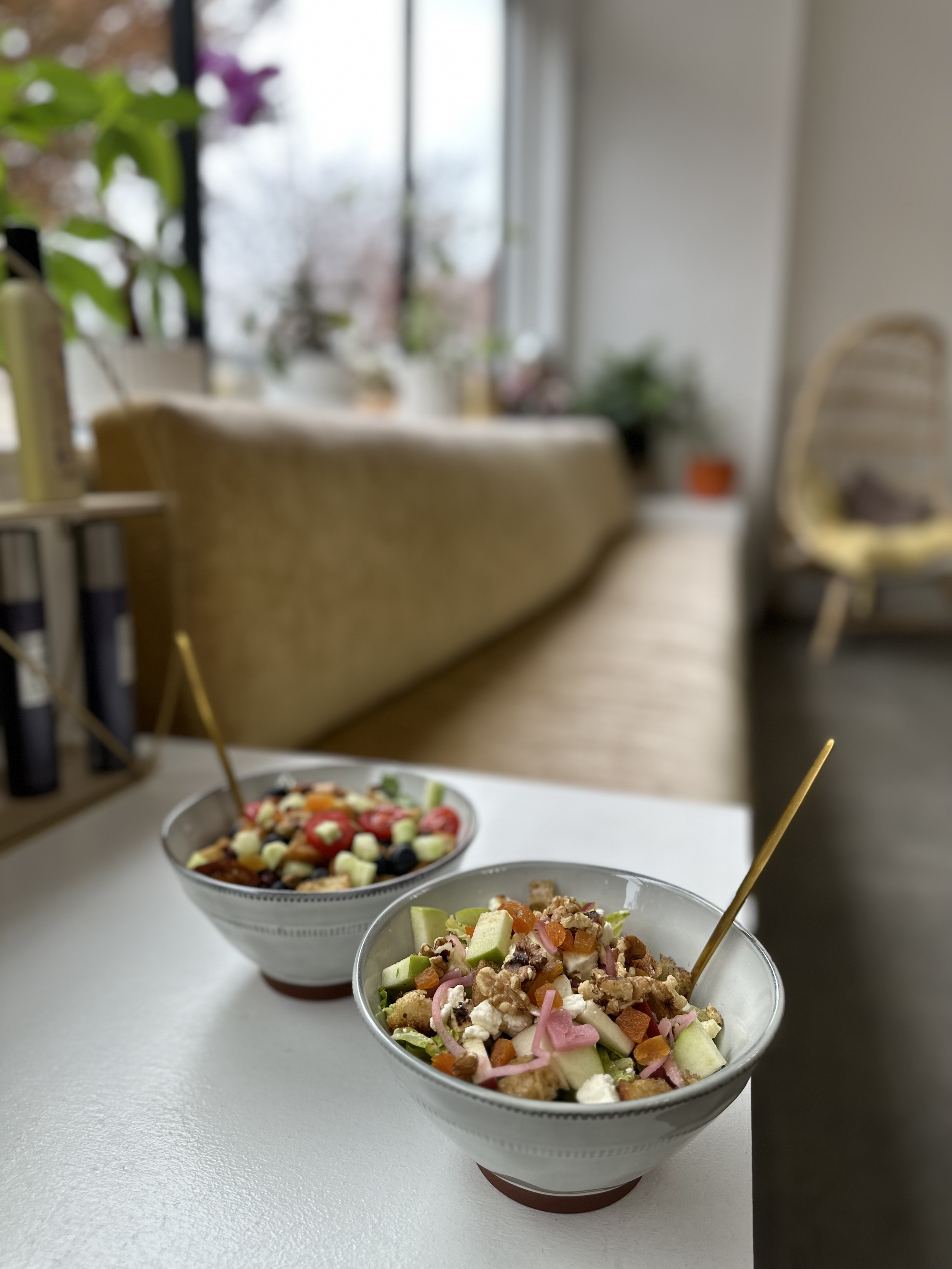 Two bowls of colorful salads with chopped vegetables, fruits, and toppings, placed on a white table in a cozy room with large windows and houseplants.