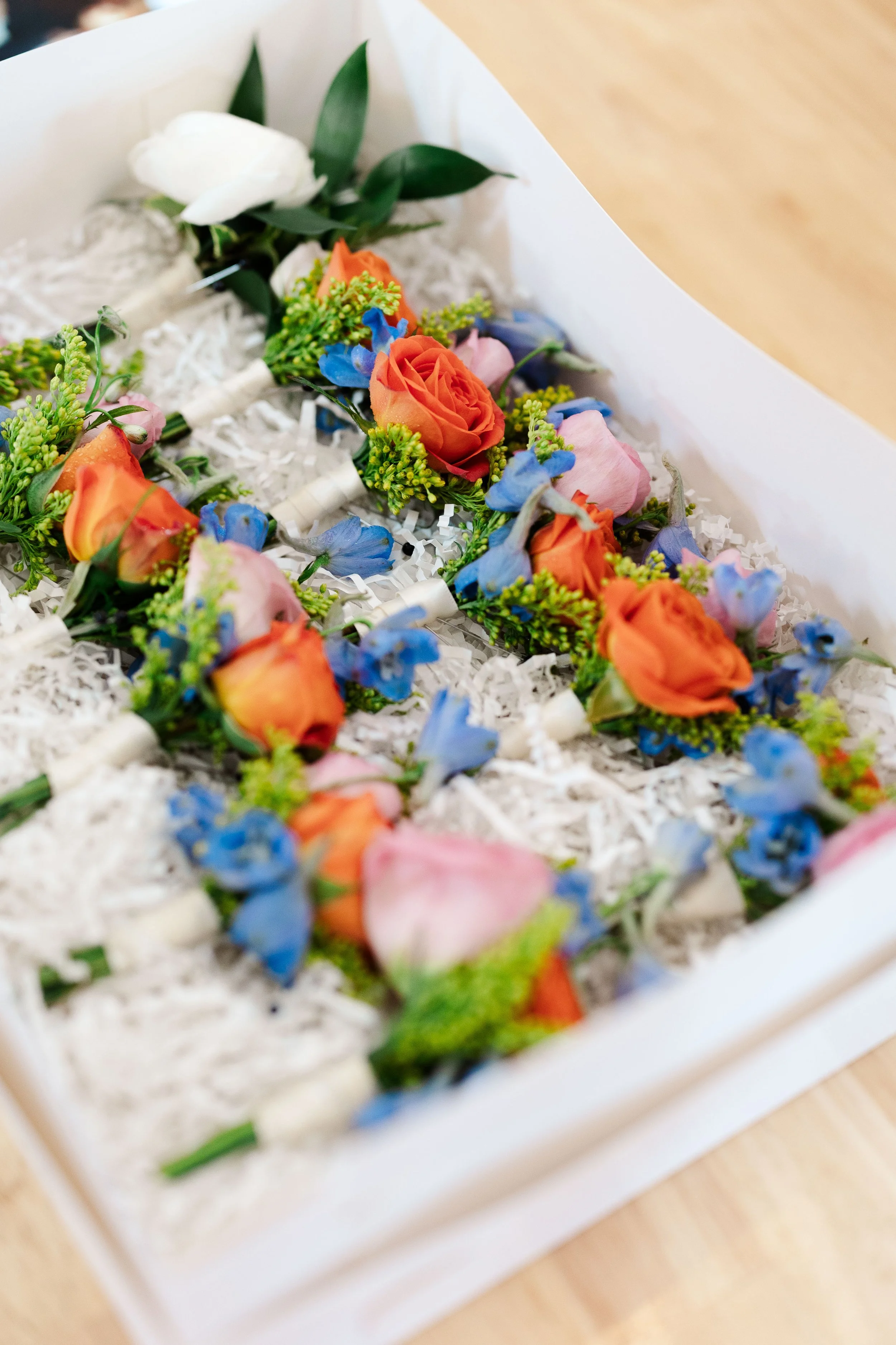 A floral boutonniere with orange roses, pink flowers, and blue accents resting on a bed of white shredded paper inside a white box.