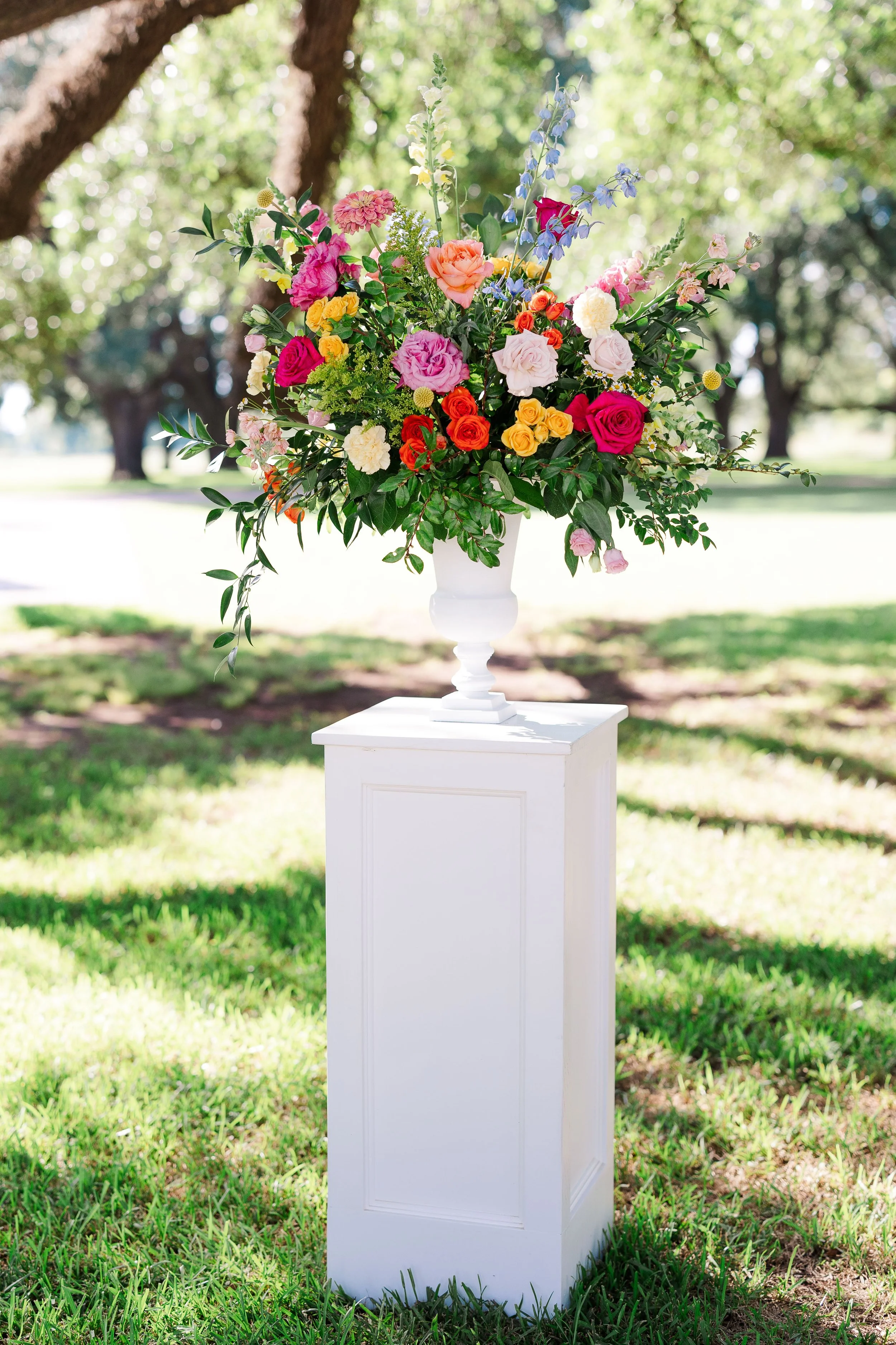 A large bouquet of colorful flowers in a white vase placed on a pedestal outdoors, with a background of trees and green grass.