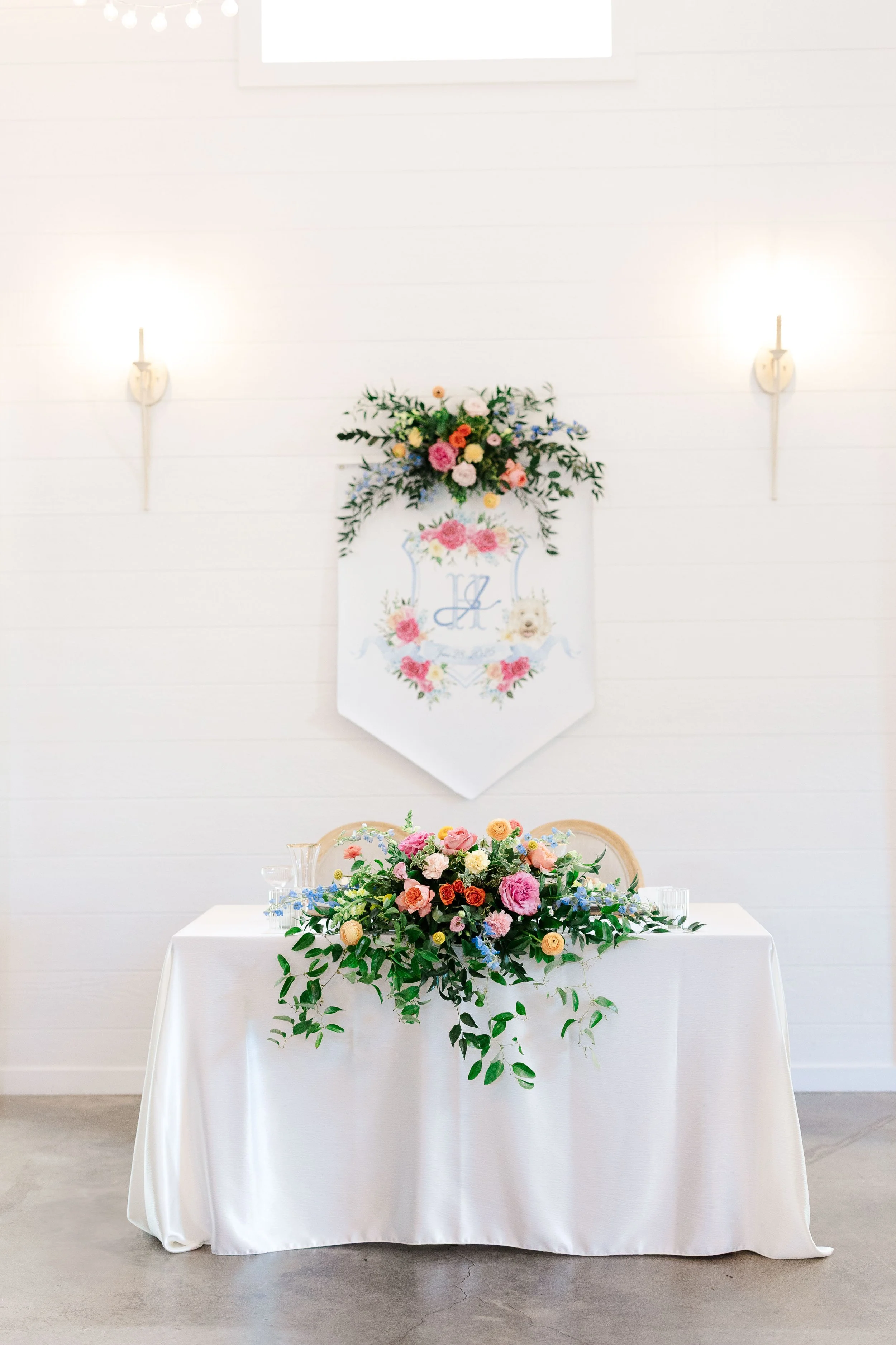 A decorated event table with a floral centerpiece and greenery, with a white tablecloth, set against a white wall with wall sconces and a floral crest banner for a celebration or wedding.