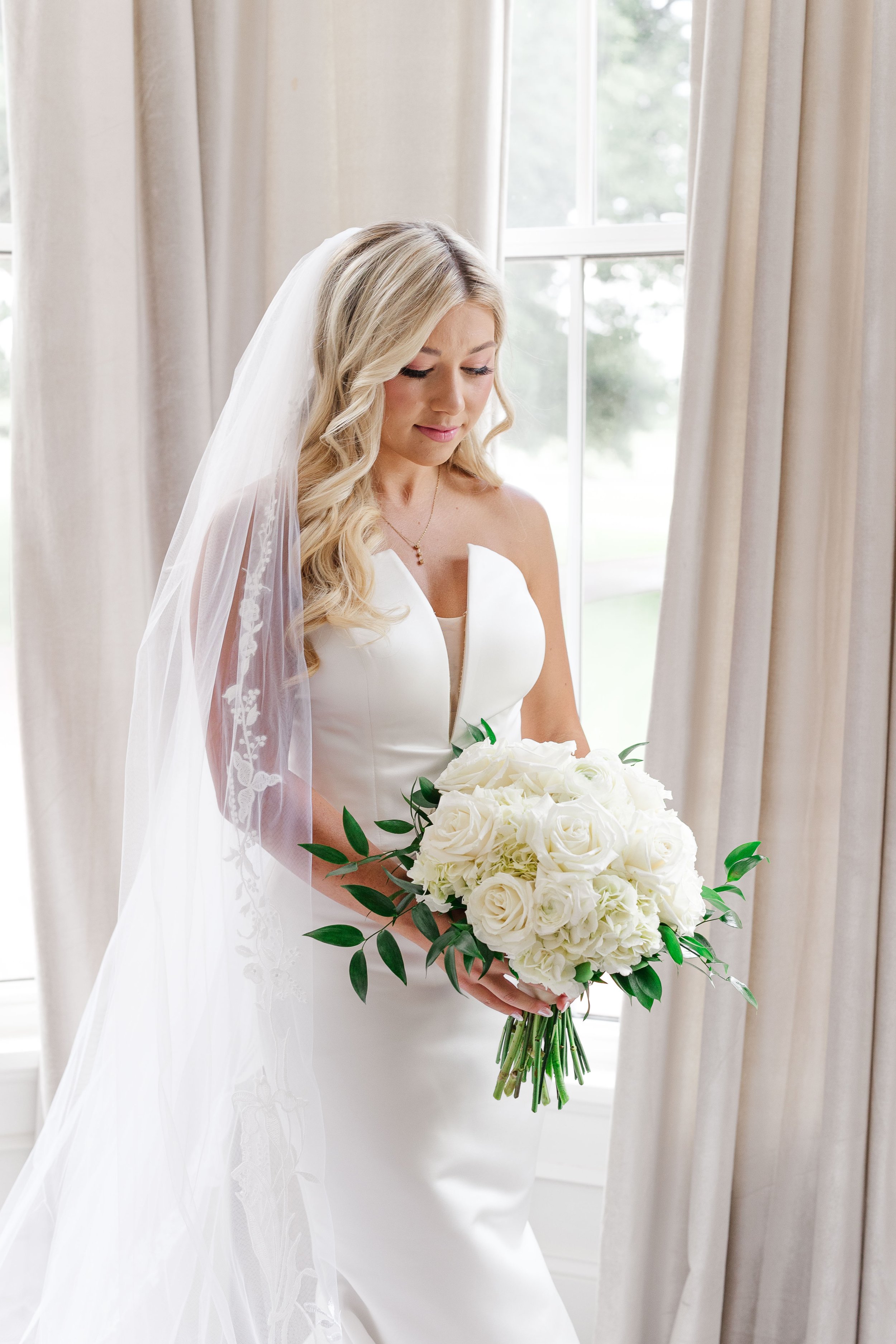 A bride with long blonde hair wearing a white wedding dress and veil, holding a large bouquet of white roses and greenery, standing by a window with beige curtains, looking down.
