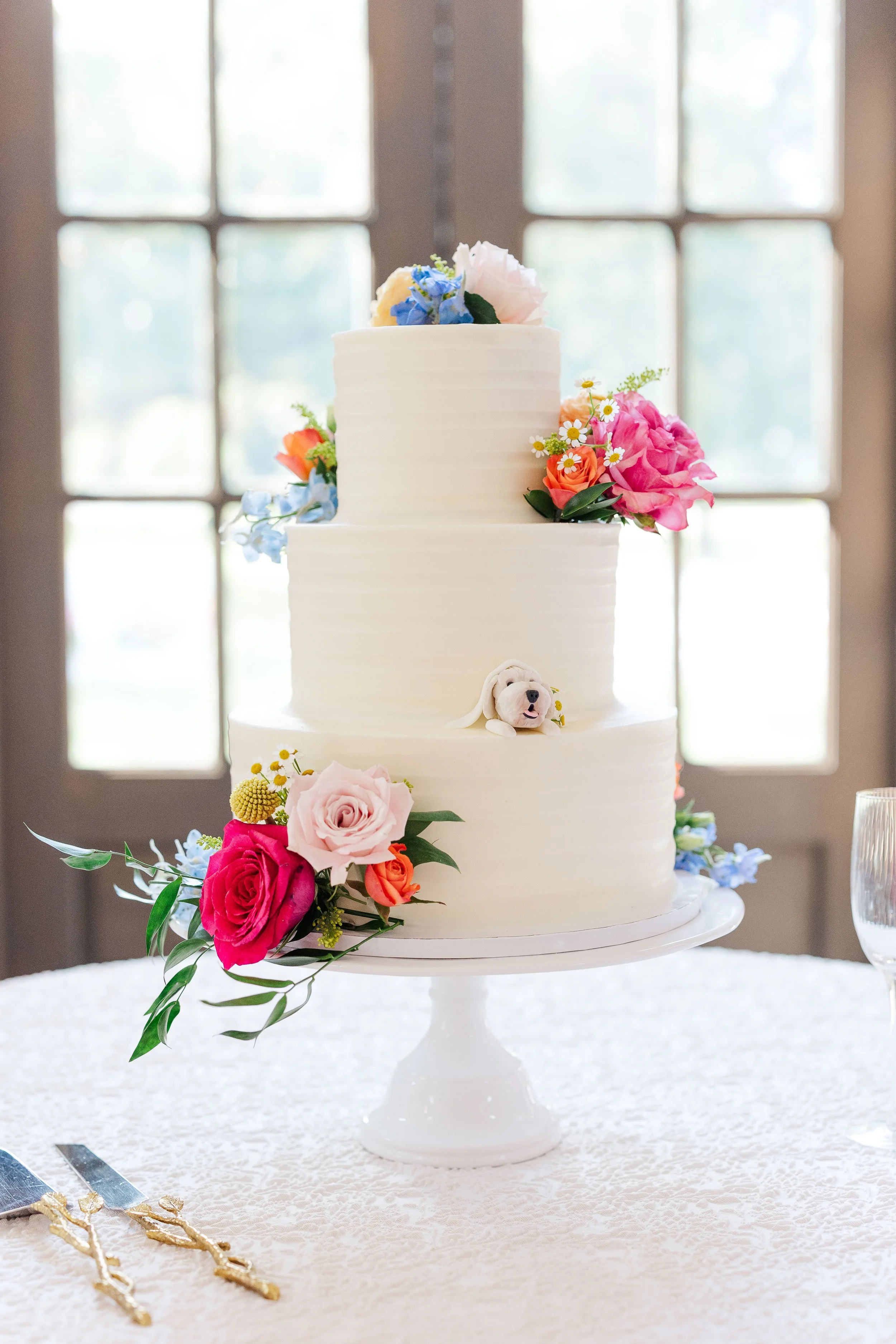 A three-tiered white wedding cake decorated with fresh flowers including pink, orange, blue, and white blossoms, with a small floral dog figurine on the middle tier, displayed on a white cake stand in front of a window.