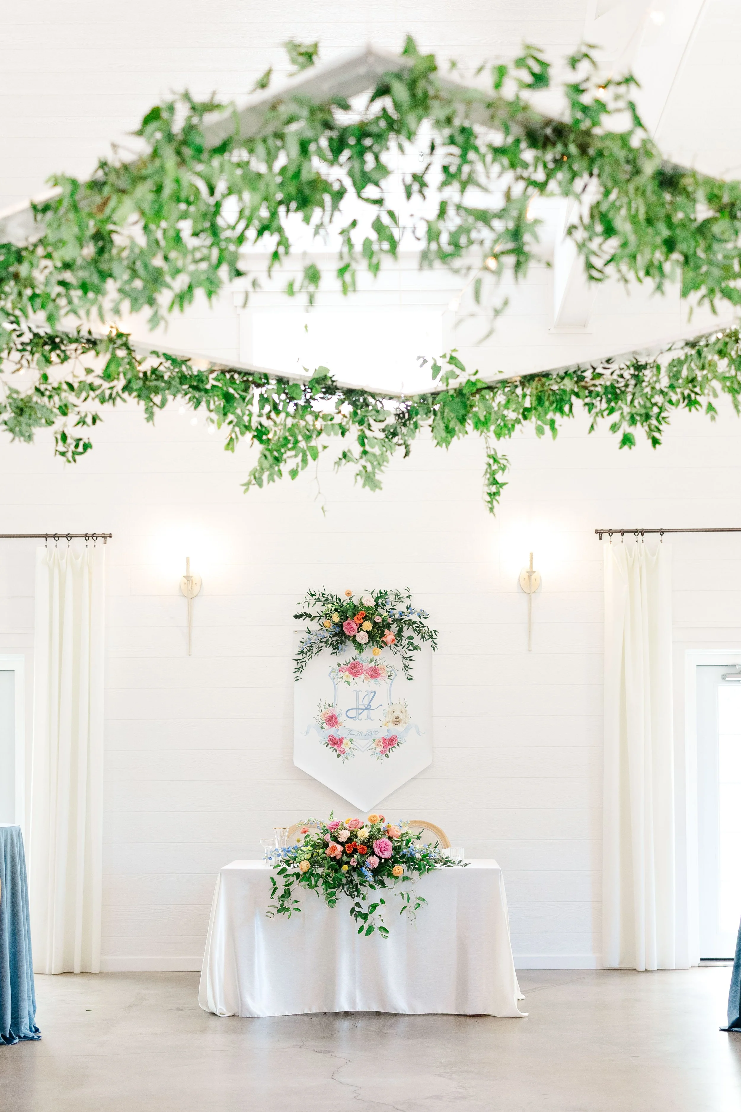 A decorated event space with a white table and a floral centerpiece, white walls, ceiling greenery, and curtains, likely set up for a celebration or wedding.