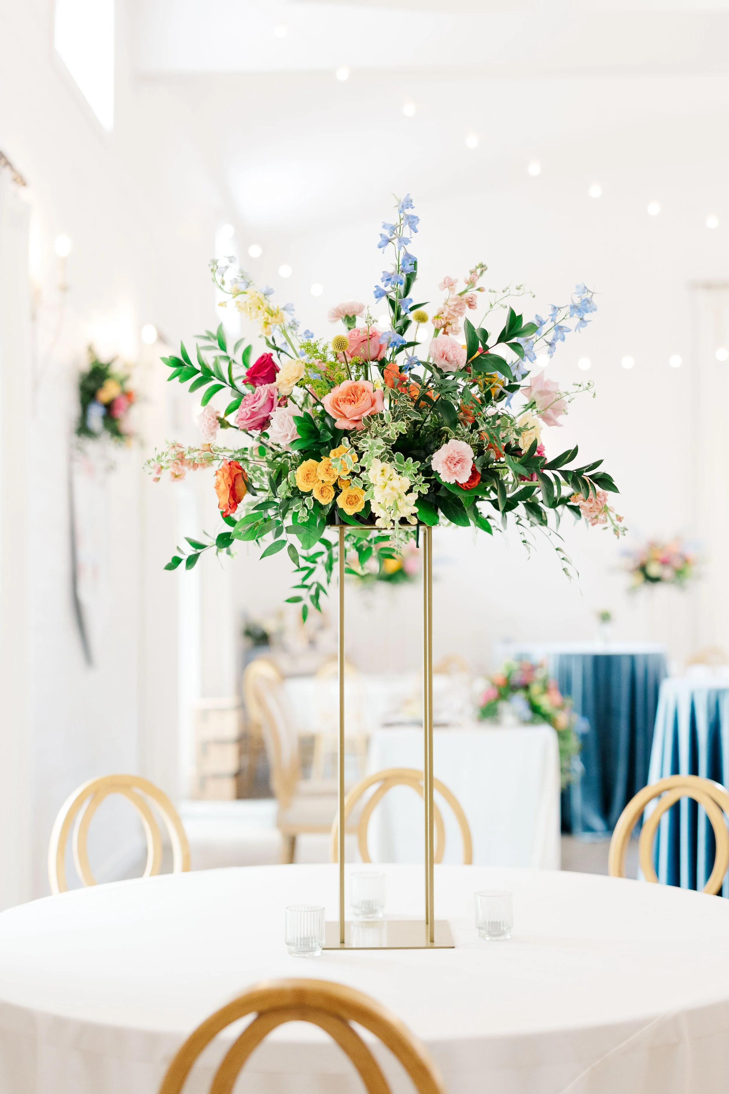 A tall flower centerpiece with pink, yellow, and blue flowers and green leaves on a white table, with a blurred background of chairs and tables, decorated with smaller flower arrangements and fairy lights.