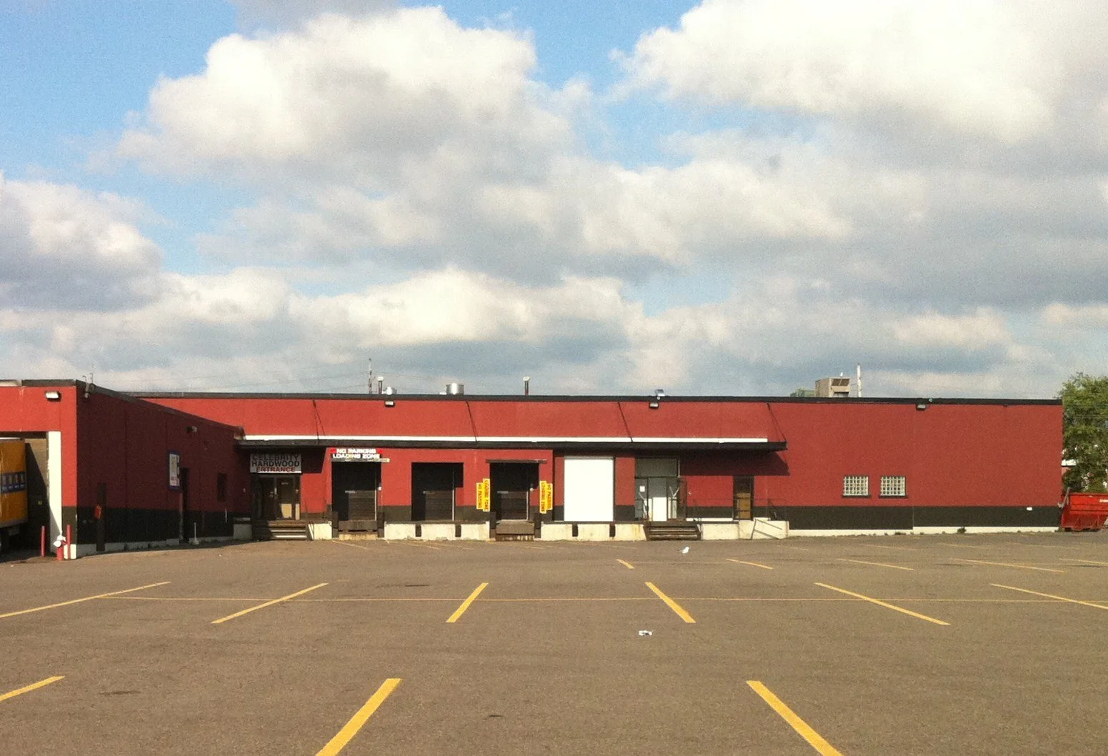 An empty parking lot in front of a red commercial building with several closed shutter doors and a few small windows.