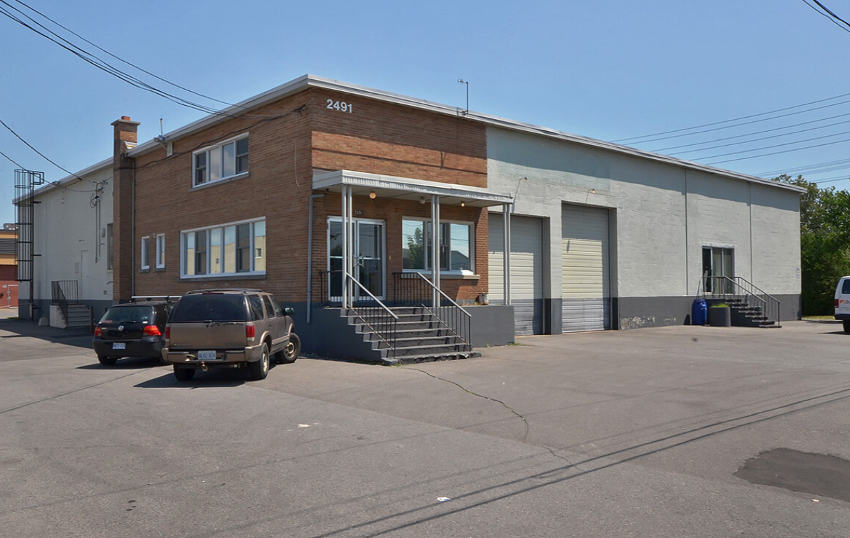 A two-story building with a brick front and gray painted side facing a parking lot with a few cars. The building has two set of stairs leading to entrances and large garage doors.